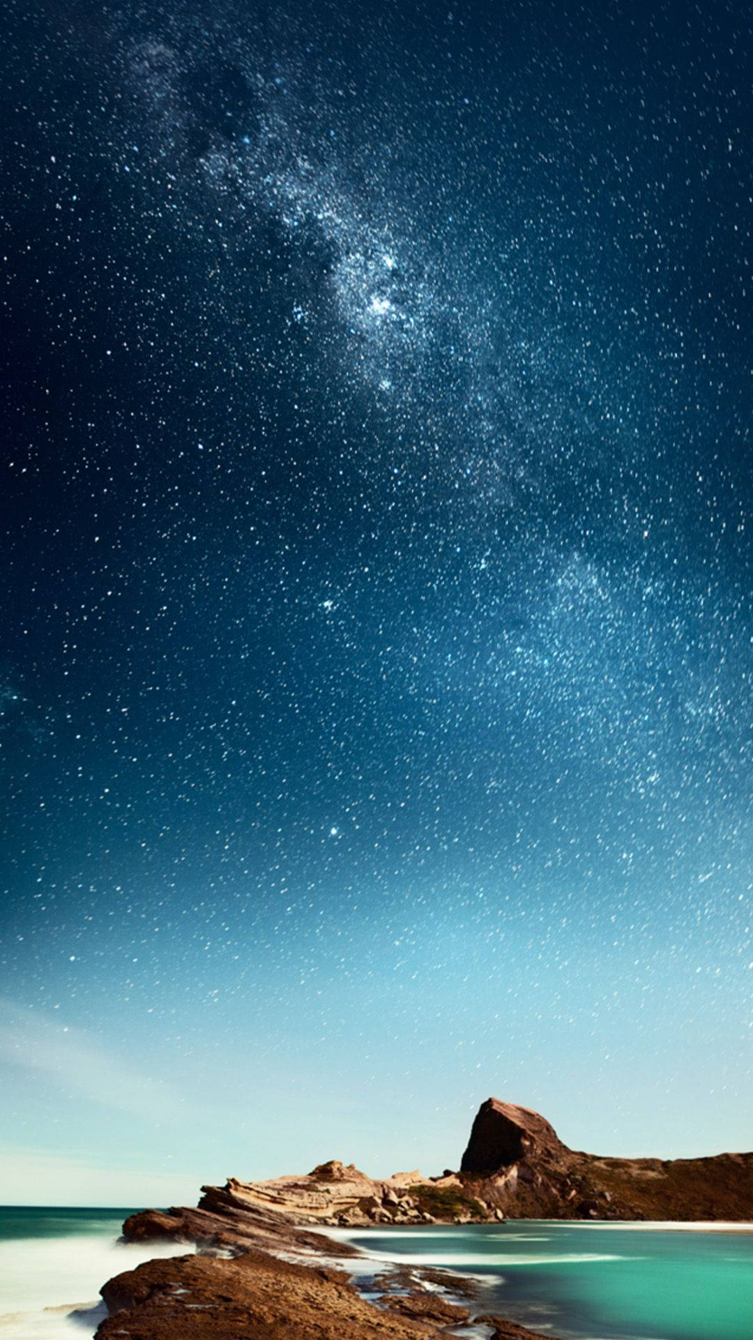 銀河灑落海灘 iPhone 壁紙 — 令人嘆為觀止的夜空，佈滿無數星辰，銀河拱跨在寧靜的碧綠海洋與崎嶇的海岸岩石之上。