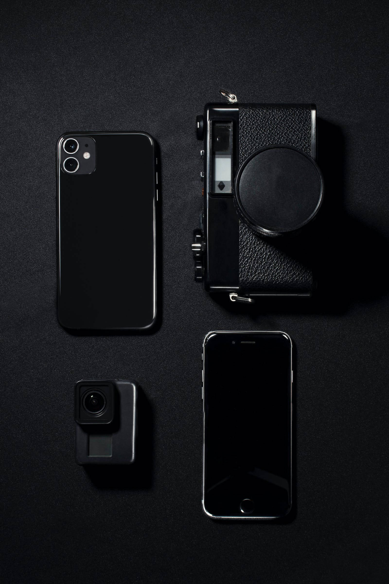 黑色美學 iPhone 電子產品收藏 壁紙 — 四款黑色科技裝置，包括智能手機和相機，整齊排列在深色紋理表面上。