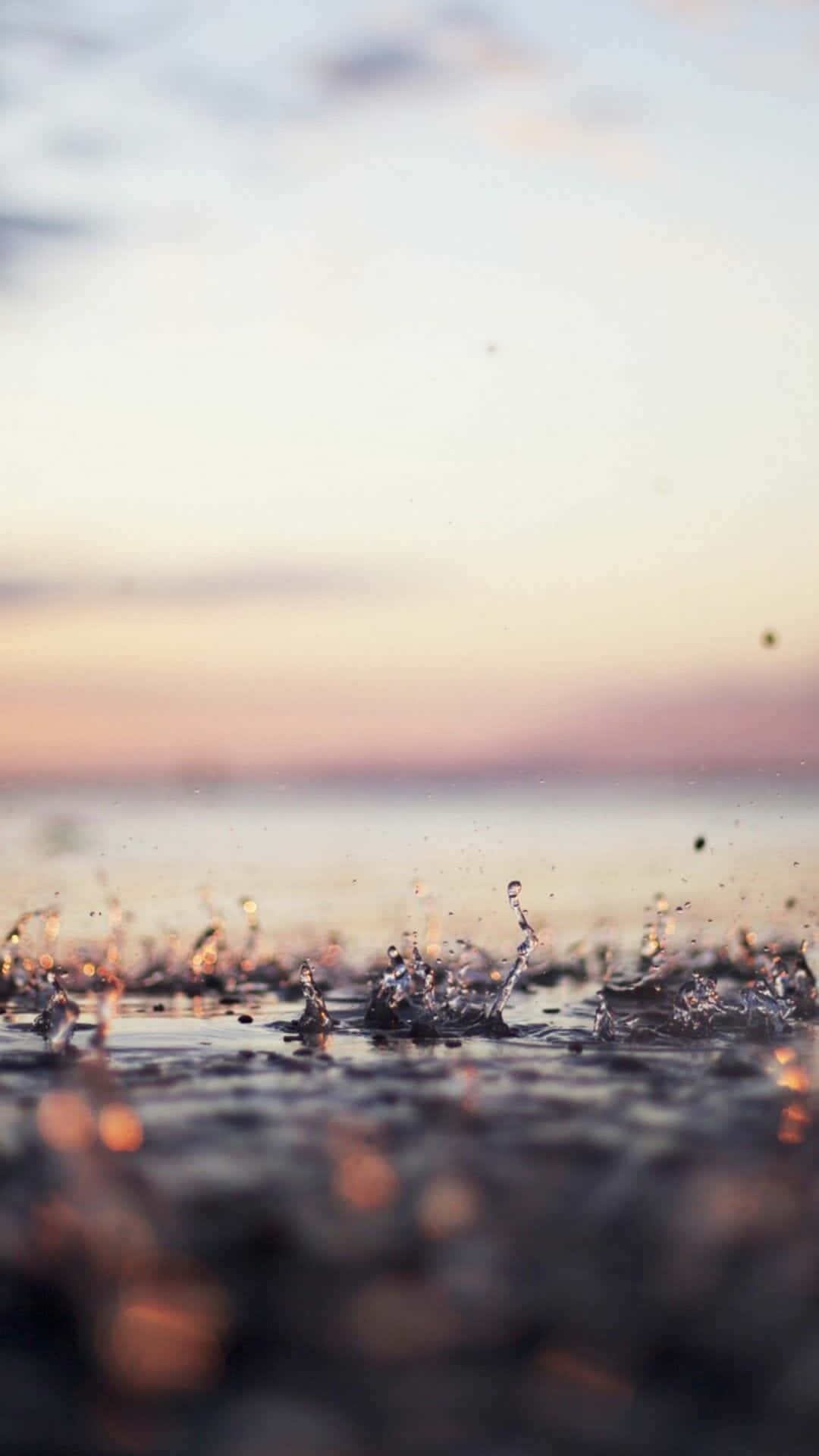 雨滴墜落 iPhone 壁紙 — 雨滴在濕漉漉的地面上戲劇性地飛濺，背景是柔和粉彩的日落天空。
