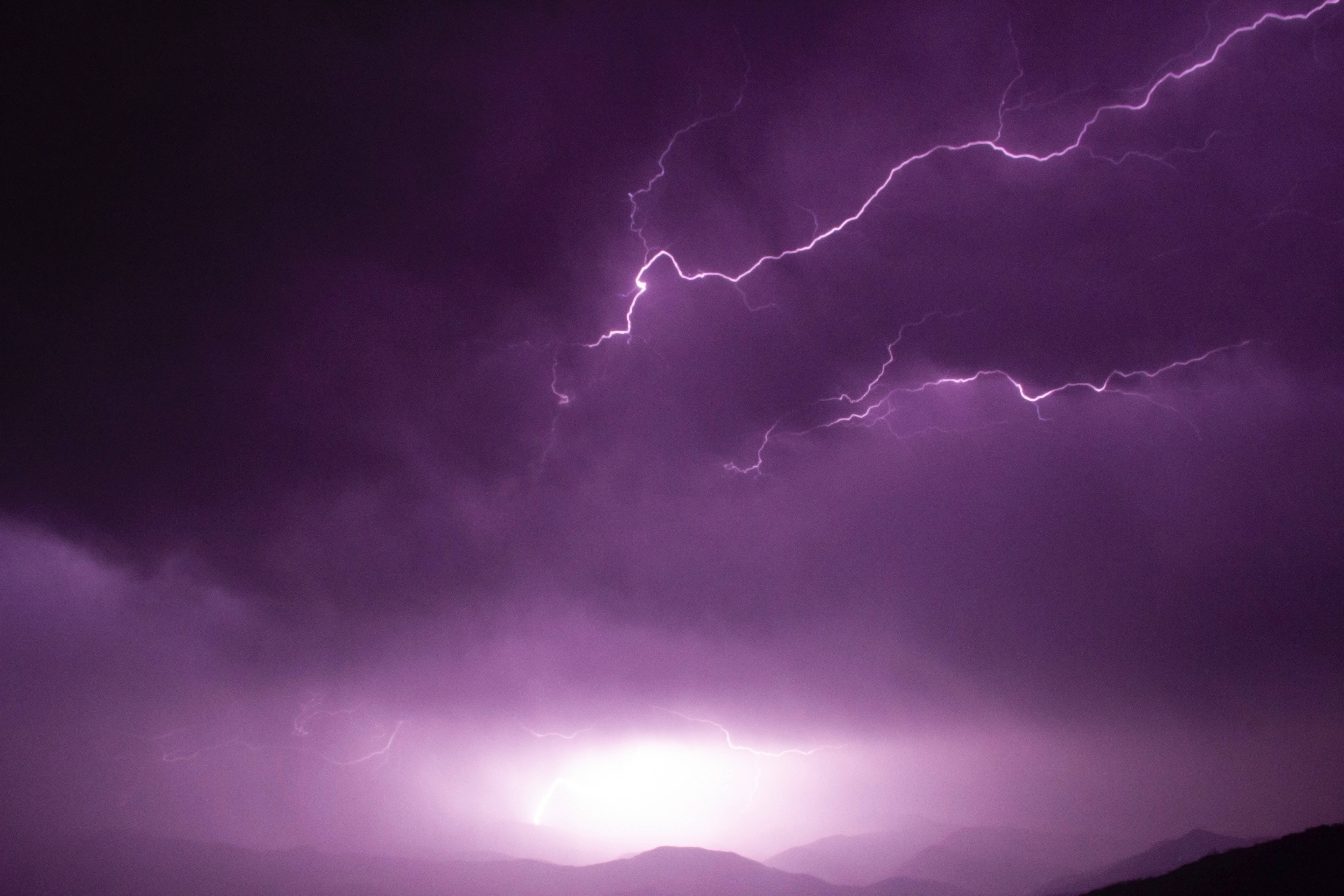 霓虹紫 iPhone 雷電天空 壁紙 — 一道戲劇性的紫色閃電在夜晚的暴風雨天空中，於剪影般的山脈上方分叉。