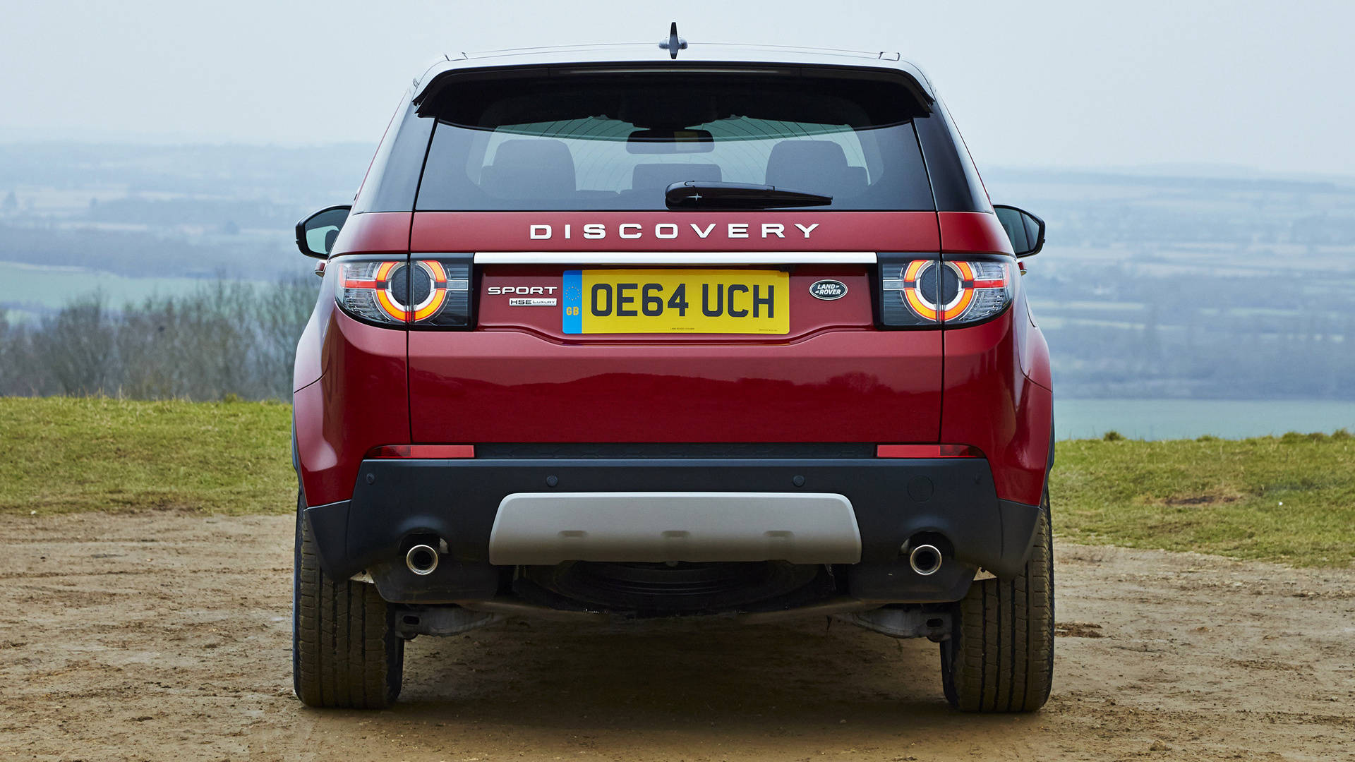 佛羅倫薩紅色發現版路虎 iPhone 主題壁紙（後視） 壁紙 — 一輛紅色 Land Rover Discovery Sport 停在一條泥路上，俯瞰著霧氣瀰漫的山谷，遠方有山丘和水面。