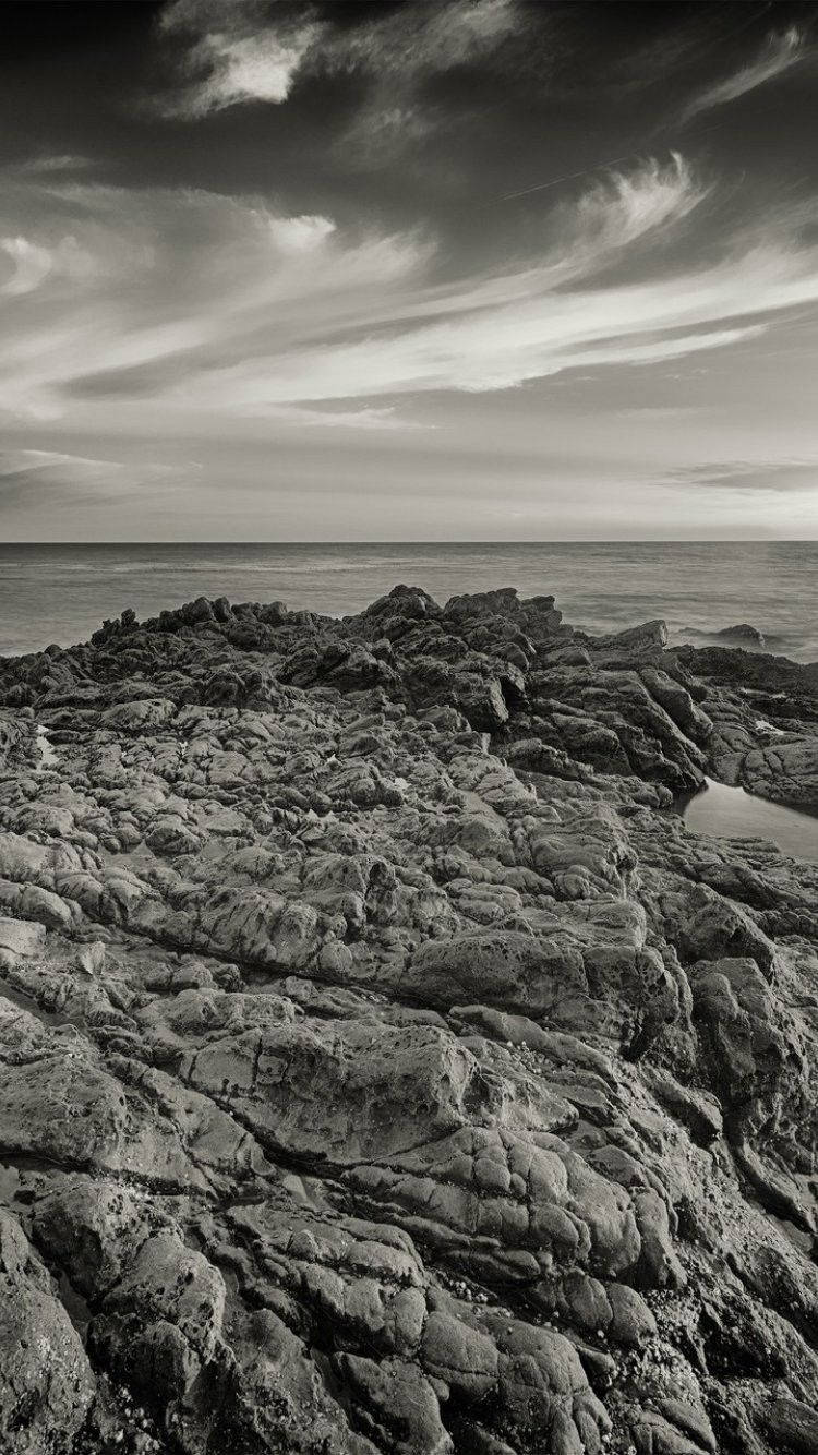 岩石海岸馬利布 iPhone 壁紙 — 單色海岸場景，紋理岩石前景，平靜海面上方動態雲層天空。