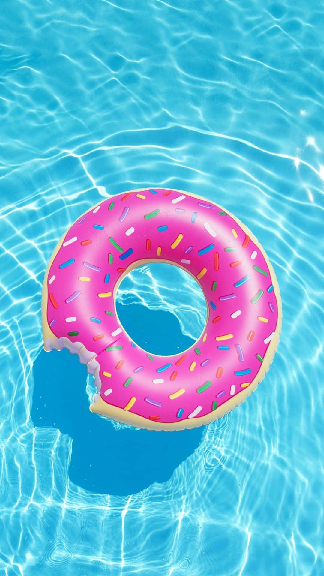 為你的夏日時尚風格增添 iPhone 壁紙！ 壁紙 — 一個粉紅色灑糖甜甜圈造型浮床，上面有咬痕，漂浮在清澈藍色泳池水中，帶有漣漪反射。