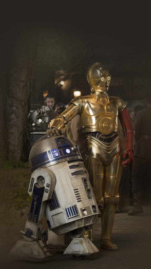 星際大戰機器人藝術 iPhone 壁紙 — 金色C-3PO和凹陷的R2-D2在夜晚昏暗的森林中並肩站立，背景有工作人員。
