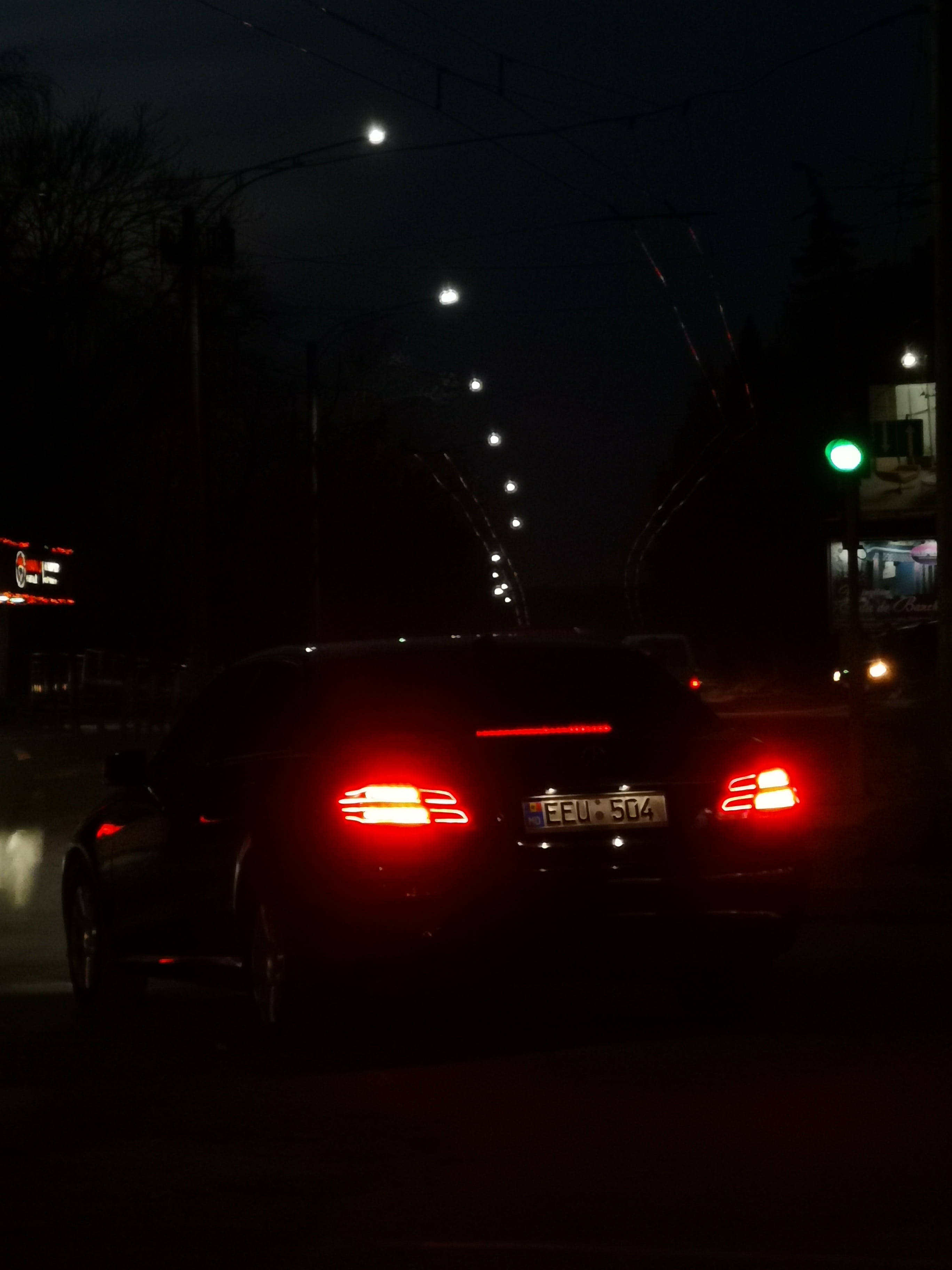 梅賽德斯 iPhone X 尾燈 壁紙 — 一輛深色轎車發光的紅色尾燈照亮了雨夜街道，街道上有一排路燈和一個綠色交通信號燈。