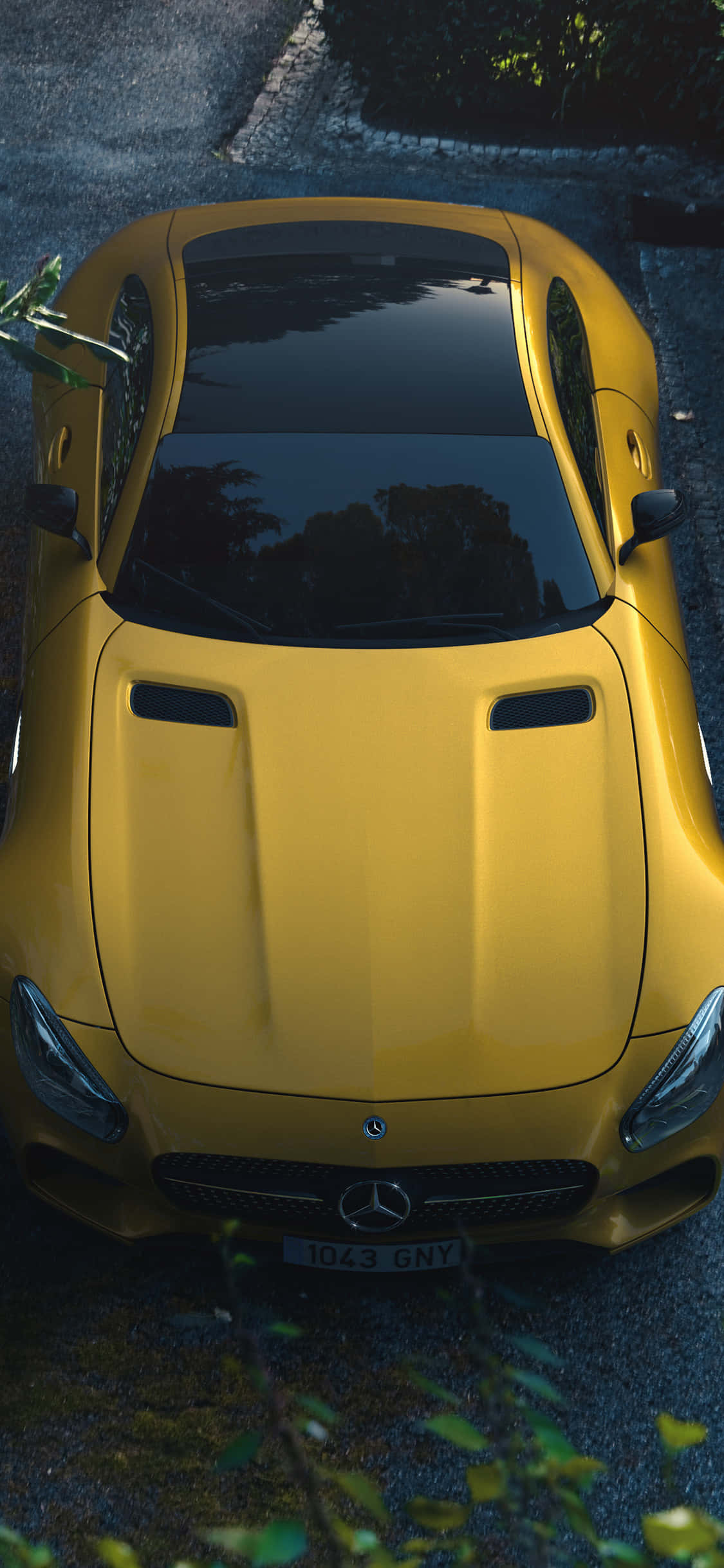 iPhone X AMG GT-R 背景黃色 AMG GT 壁紙 — 從上方俯瞰一輛充滿活力的黃色 Mercedes-AMG GT 跑車停泊在鵝卵石街道上，周圍有綠色植物。