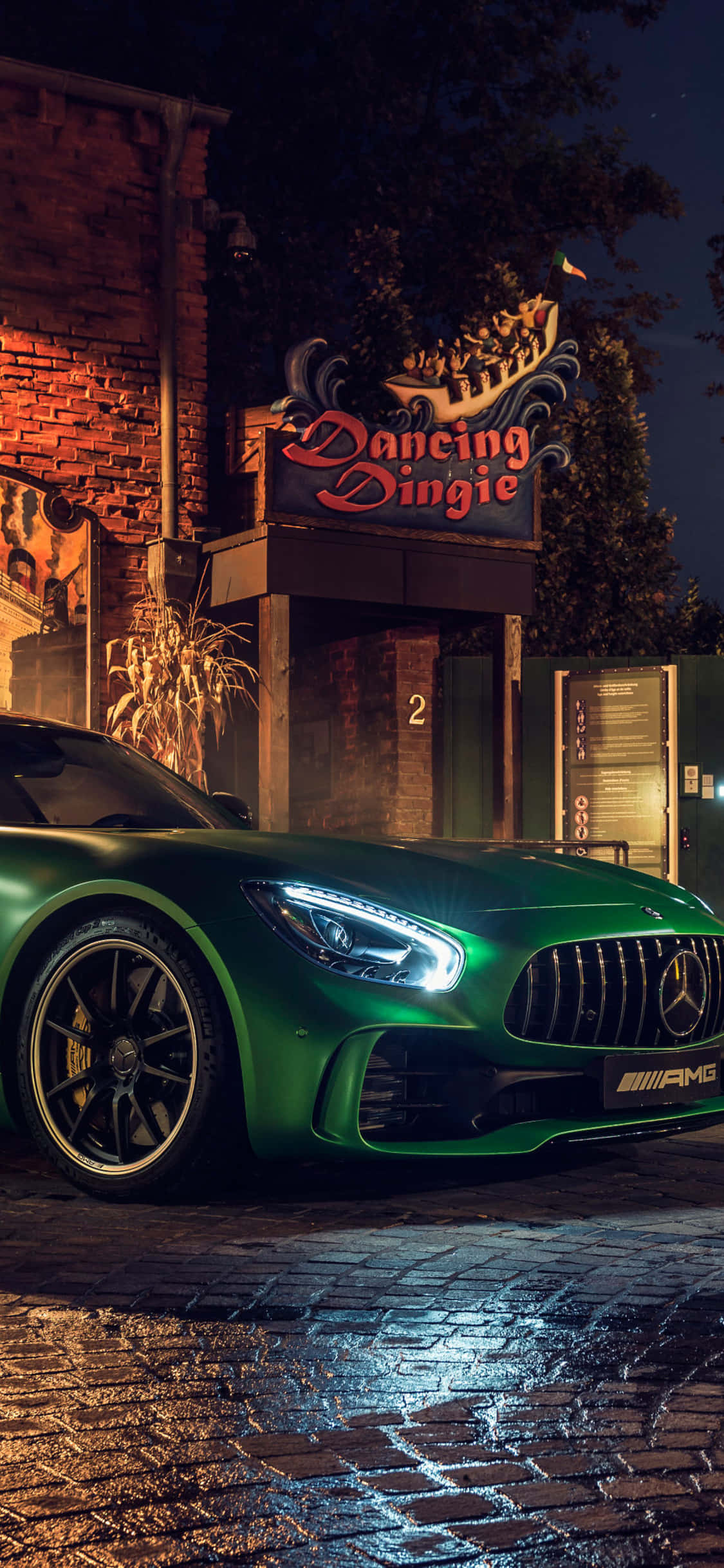 iPhone X AMG GT-R 背景與綠色 AMG GT 壁紙 — 一輛光亮的綠色AMG跑車在霓虹燈下閃耀，停在夜間濕漉漉的鵝卵石街道上，靠近一棟帶有 'Dancing Dingie' 標誌的磚砌建築。