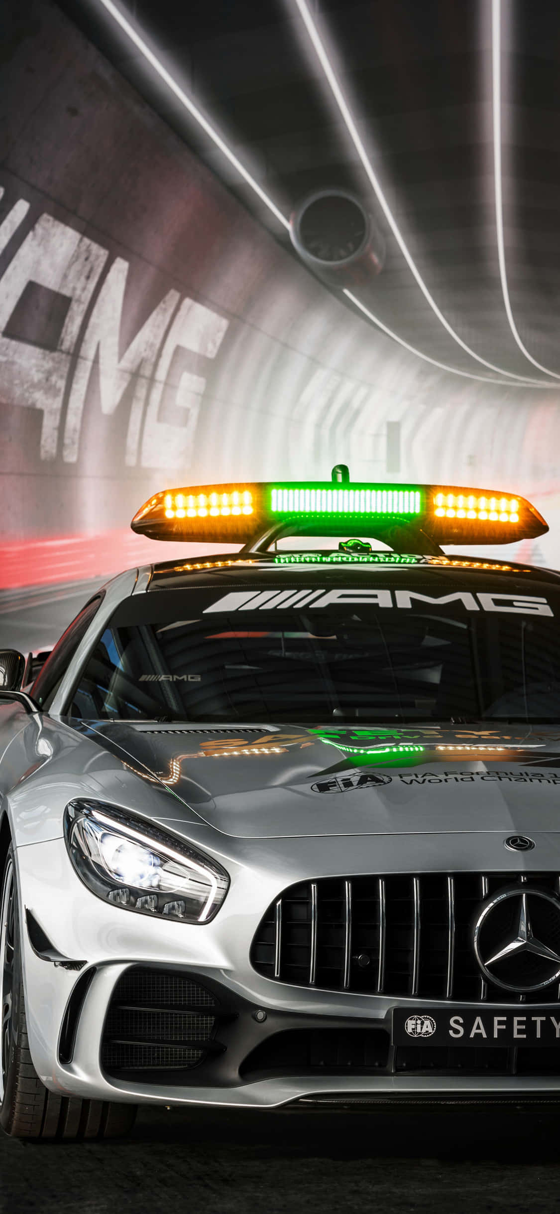 iPhone X AMG GT-R 背景 白色 AMG GT 壁紙 — 一輛銀色 Mercedes-AMG 安全車，帶有閃爍的橙色和綠色燈光，在牆上有 'AMG' 塗鴉的隧道中疾馳。