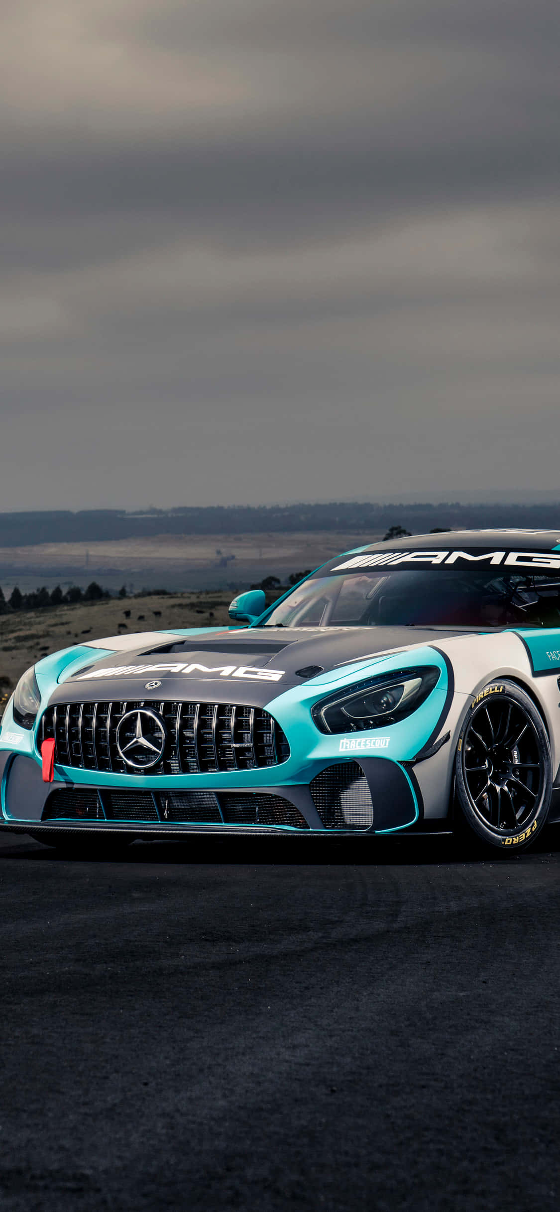 iPhone X AMG GT-R 背景：雙色 AMG GT4 駛過陰雲密布天際 壁紙 — 一架時尚的藍綠色和白色平治AMG賽車，在陰沉多雲的天空下，停泊在漆黑的瀝青賽道上。