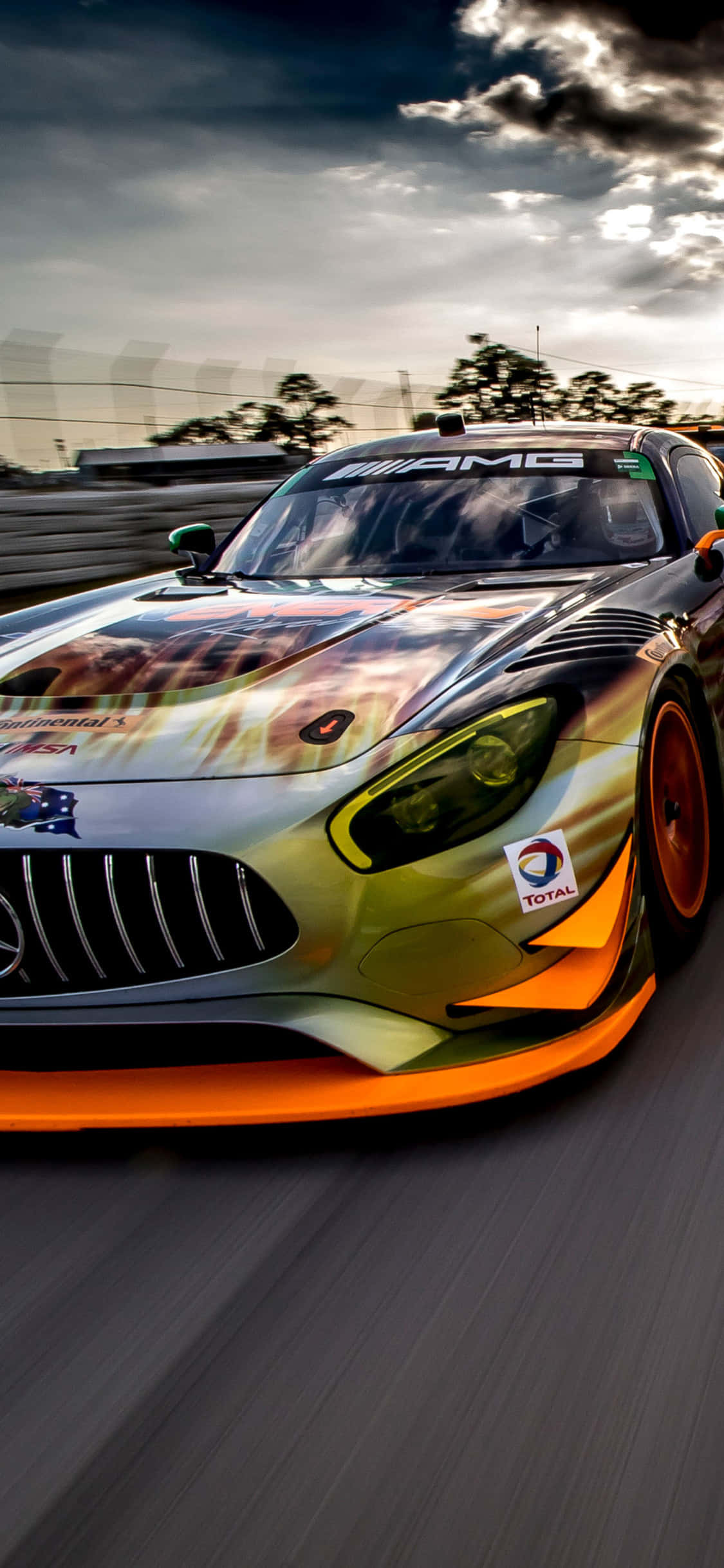 iPhone X AMG GT-R背景搭配獵豹紋理 壁紙 — 一輛帶有火熱橙色和金色塗裝的Mercedes-AMG GT3賽車，在戲劇性的多雲天空下沿賽道飛馳。