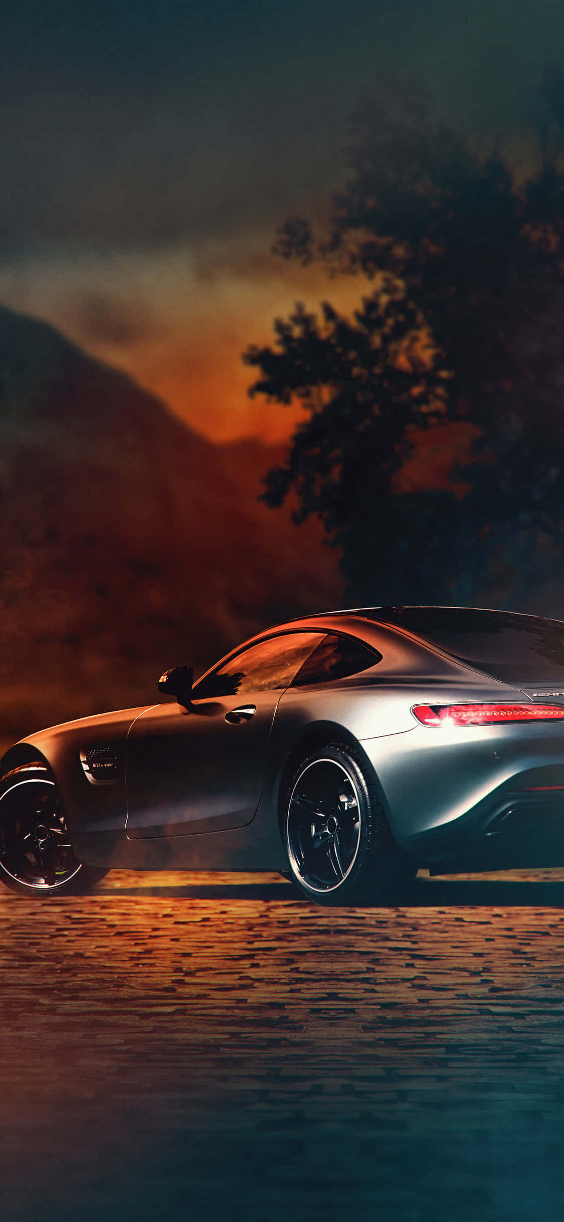 iPhone X AMG GT-R 背景與銀色 AMG GT C 壁紙 — 一輛時尚的銀色跑車在鵝卵石路上行駛，天空是火紅的日落，襯托出樹木和山脈的剪影。