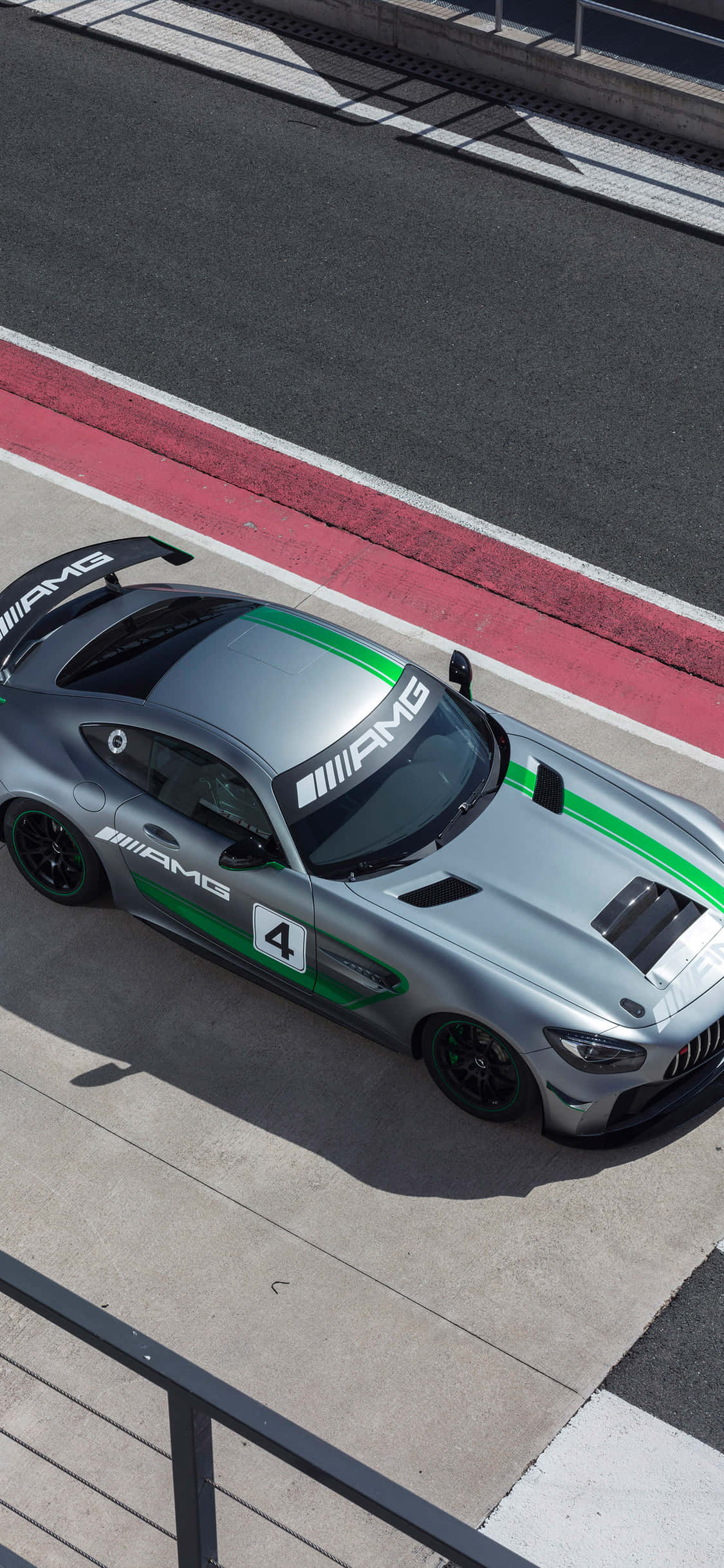 iPhone X AMG GT-R背景與梅賽德斯-AMG GT4 壁紙 — 一輛帶有綠色條紋的時尚銀色AMG賽車在明亮的日光下沿著紅色路緣的賽道飛馳。