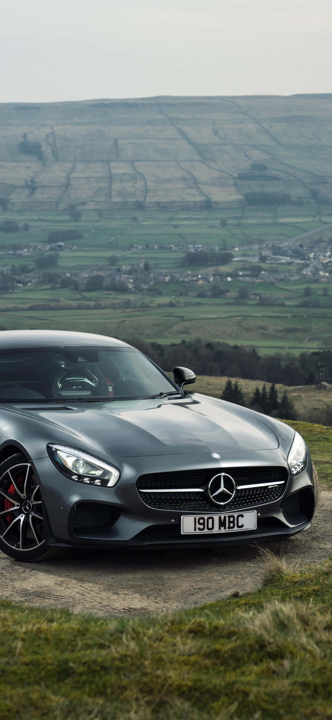 iPhone X AMG GT-R 背景山景 AMG GT 壁紙 — 一架時尚的灰色Mercedes-AMG GT停泊在一個草坡上，俯瞰著一個廣闊的鄉村山谷，山谷中有拼布般的田野和遠處的村莊，天空多雲。