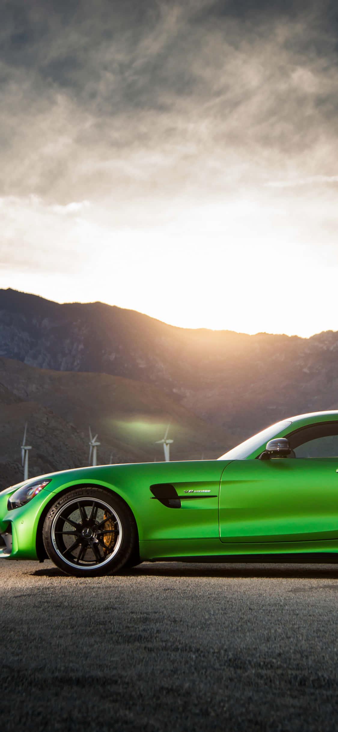 iPhone X AMG GT-R 背景：綠色梅賽德斯-AMG GT 側面車頭 壁紙 — 一架充滿活力的綠色跑車停在瀝青路上，背景有風力發電機和山脈，天空呈現金黃色的日落。
