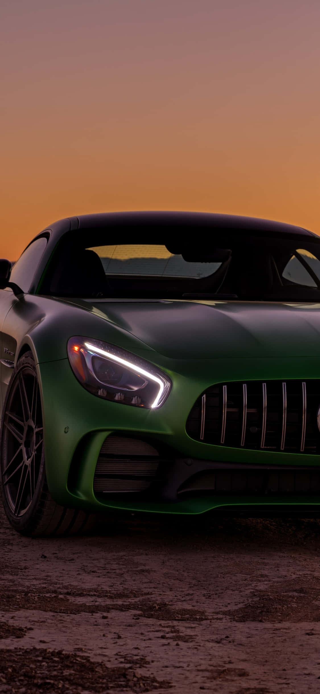 iPhone X AMG GT-R 背景：綠色梅賽德斯-AMG 前燈亮起 壁紙 — 一輛時尚的綠色跑車在溫暖的橙色日落天空下閃耀，車頭燈發出光芒。