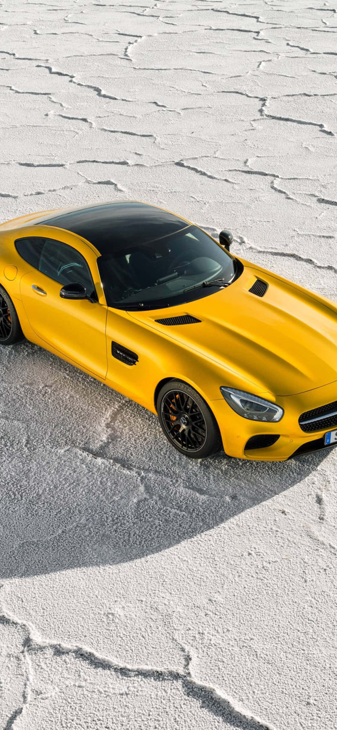 iPhone X AMG GT-R背景與梅賽德斯-AMG GT 壁紙 — 一輛充滿活力的黃色跑車帶有黑色裝飾，停在龜裂的白色鹽灘上，沐浴在明亮的日光下。
