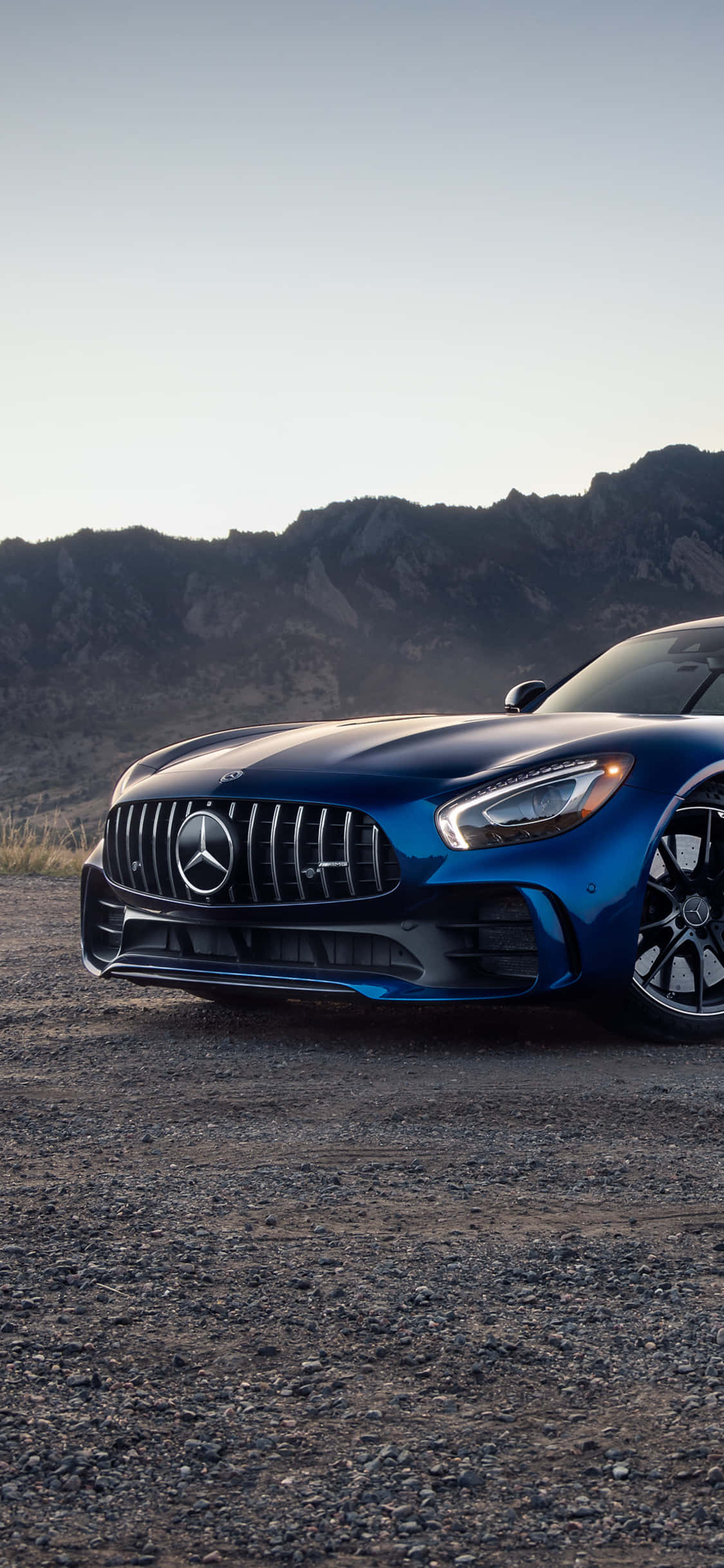 iPhone X AMG GT-R背景與梅賽德斯-AMG GT 壁紙 — 一架時尚的藍色 Mercedes-AMG GT 停泊在碎石上，背景是崎嶇的山脈，天空柔和。