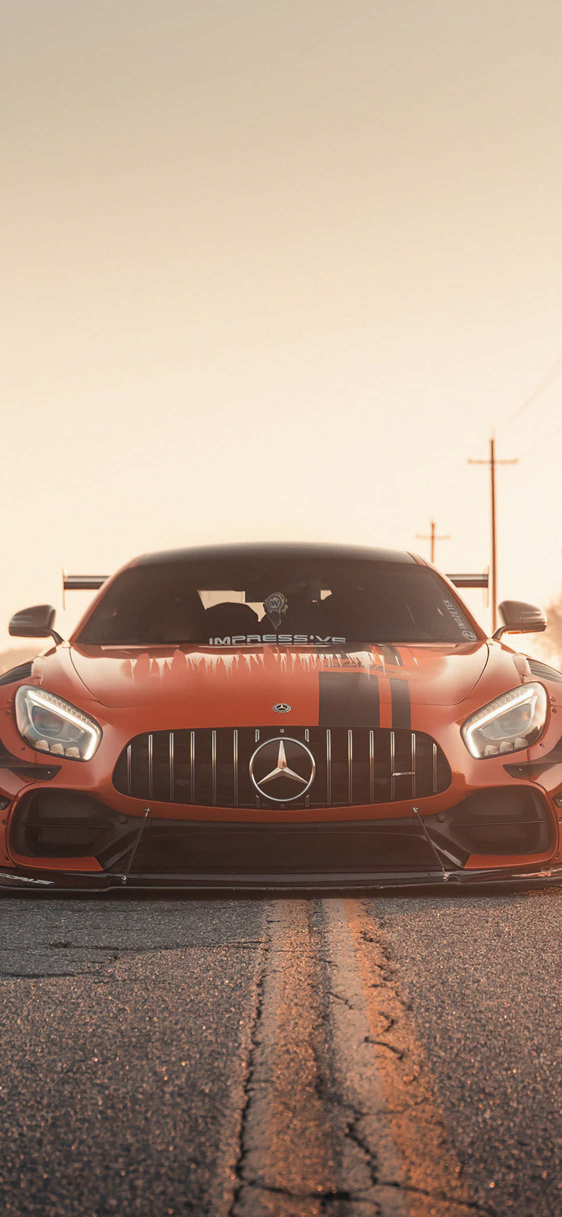 iPhone X AMG GT-R 背景橙色前臉 AMG GT 壁紙 — 一架火橙色Mercedes-AMG GT賽車在龜裂的瀝青路上向前行駛，背景是朦朧的金色日落。