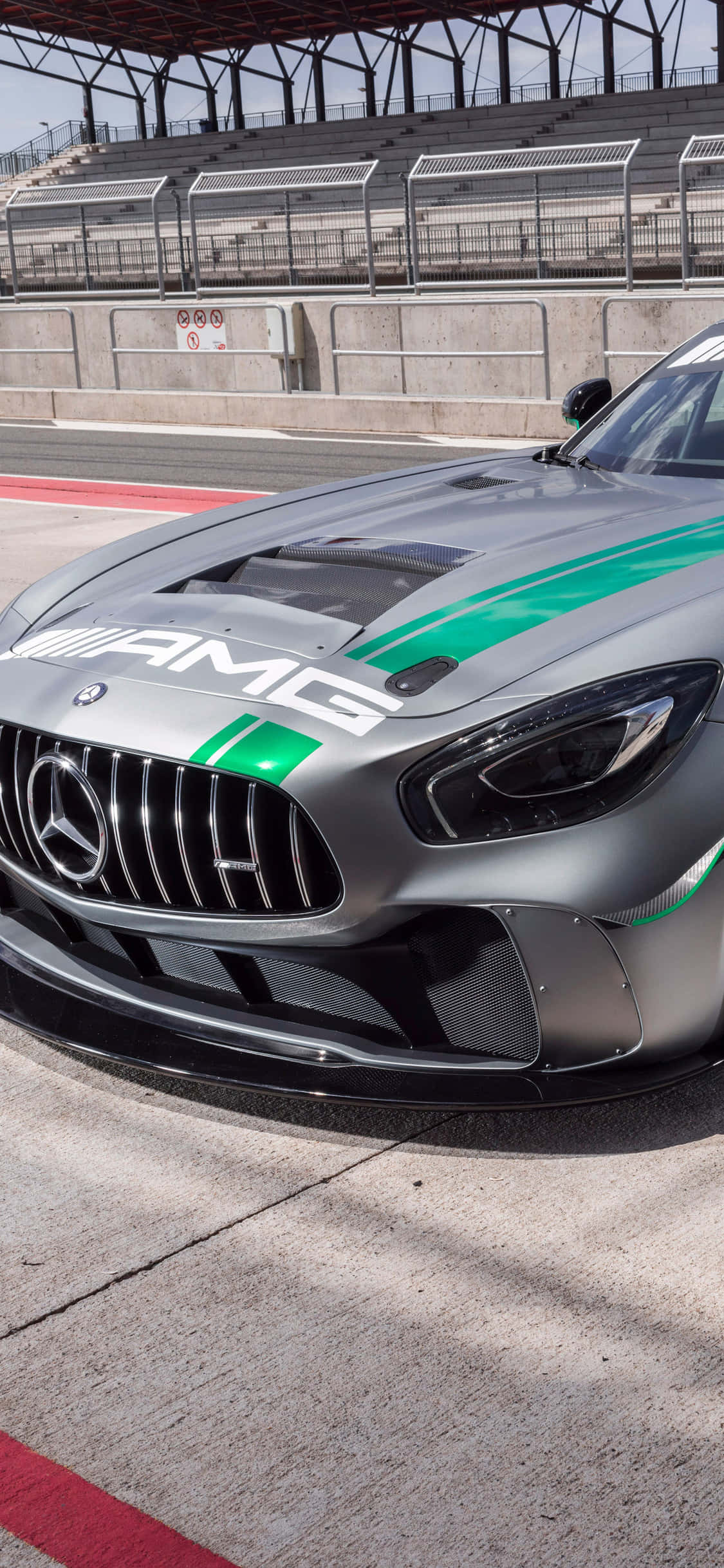 iPhone X AMG GT-R 背景：銀色 AMG GT4 賽道停泊 壁紙 — 一架時尚的銀色AMG賽車，帶有綠色條紋，停在混凝土賽道上，在明亮的日光下。