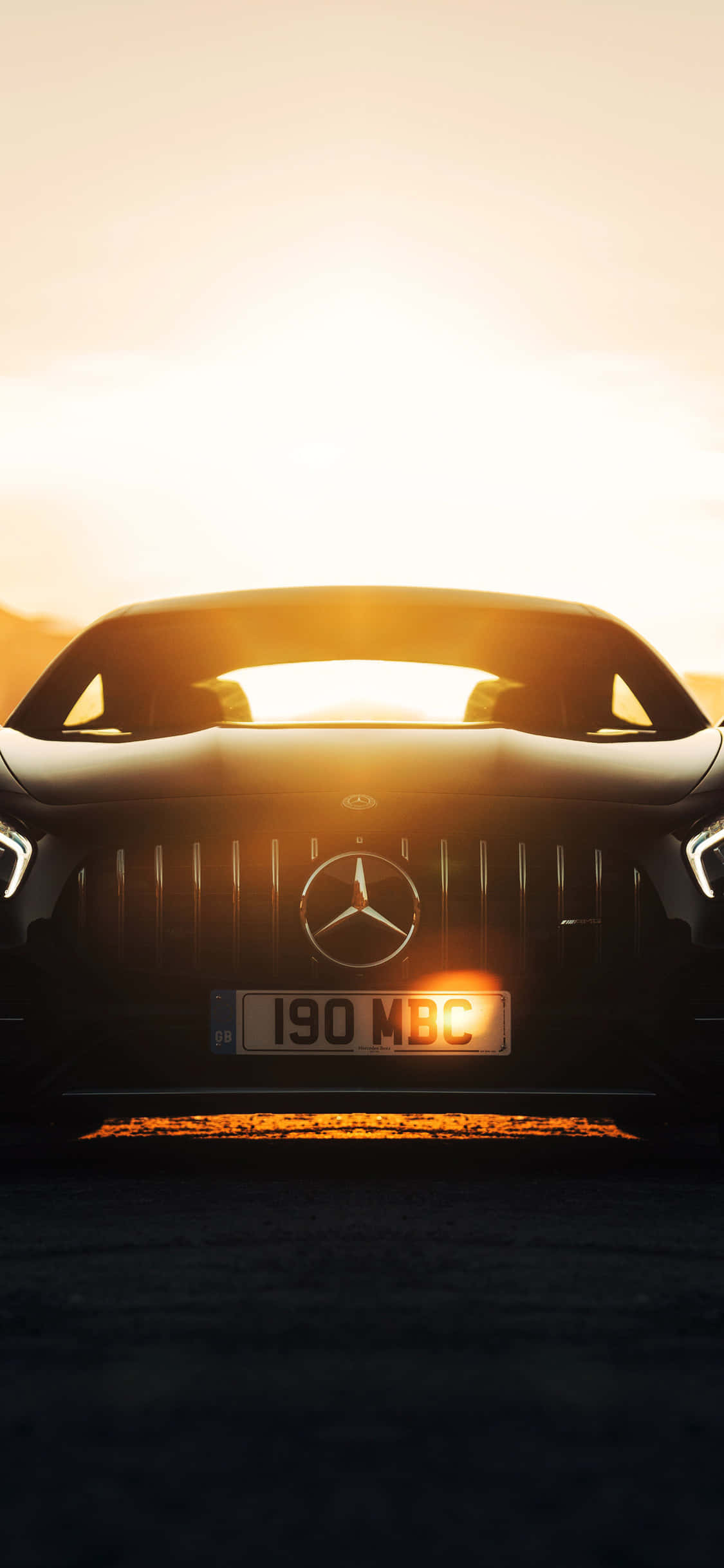 iPhone X AMG GT-R 背景 日光灑落 壁紙 — 一架時尚的黑色梅賽德斯跑車面向鏡頭，金色夕陽的光芒在其格柵和擋風玻璃後方閃耀。