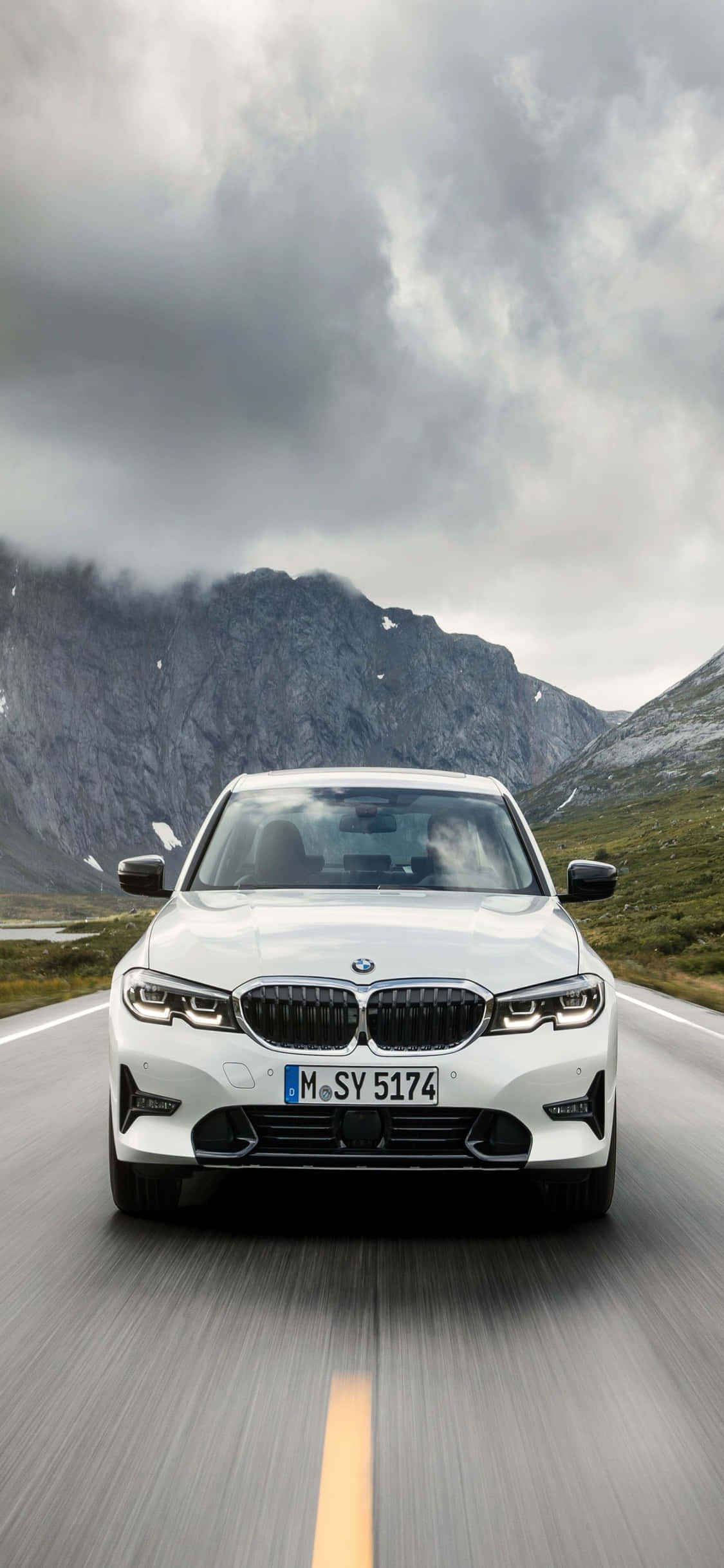 融合效能與時尚風格 – iPhone X 與 BMW 壁紙 — 一輛白色BMW轎車在戲劇性的多雲天空下，背景是崎嶇的山峰，沿著山路疾馳。