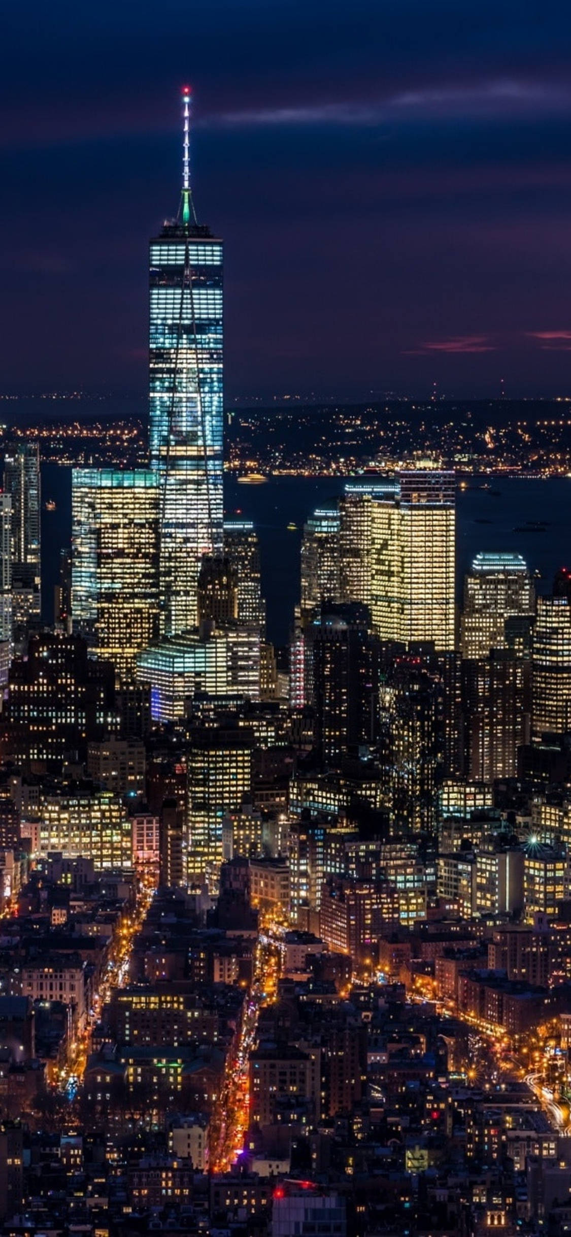 紐約市 iPhone X 夜景 壁紙 — 一座高聳的摩天大樓在深紫色的夜空下閃爍著藍光，下方是密集的城市景觀，建築物燈火通明，街道閃爍發光。