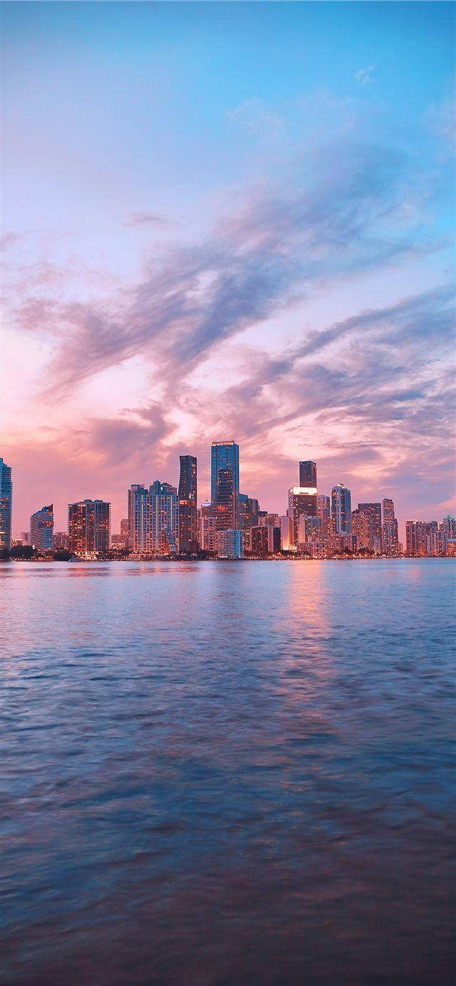 iPhone X 風格邁阿密建築風景 壁紙 — 日落時分的城市天際線，粉紅色和藍色的雲彩倒映在平靜的水面上。