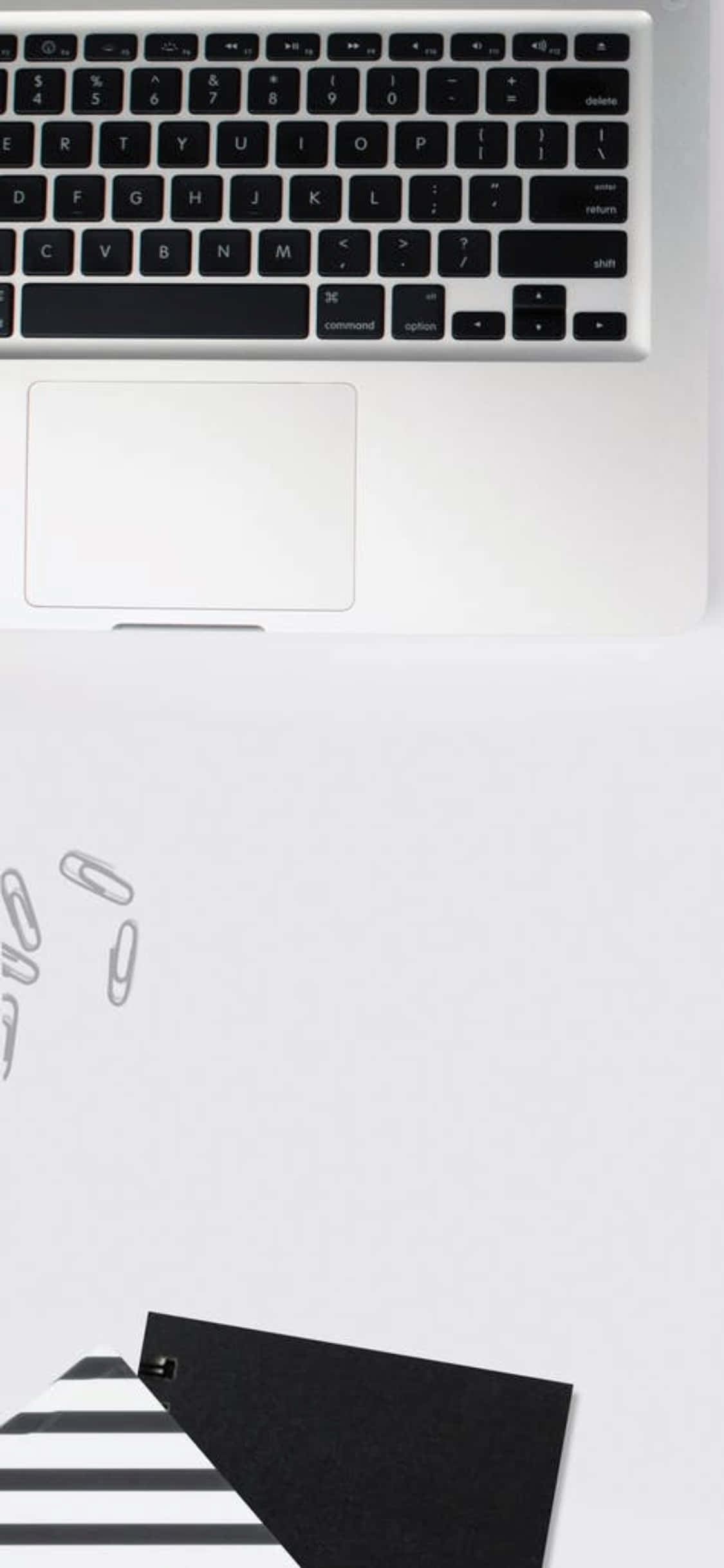 iPhone X 書桌背景：筆電與紙夾 壁紙 — 白色書桌上銀色手提電腦鍵盤和觸控板的俯視圖，散落著萬字夾和一個黑白條紋文件夾。