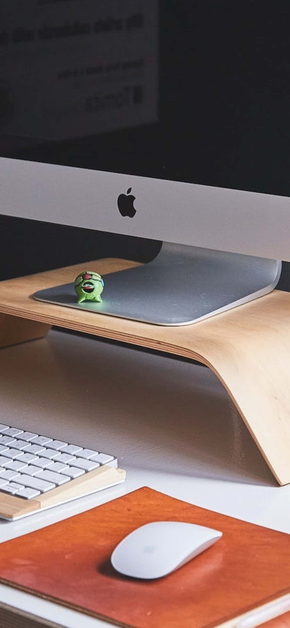 iPhone X 桌面背景：MacBook Pro 壁紙 — 一個簡約的桌面設置，包括 Apple iMac、白色鍵盤和滑鼠放在木製支架上，並有一個綠色小公仔。