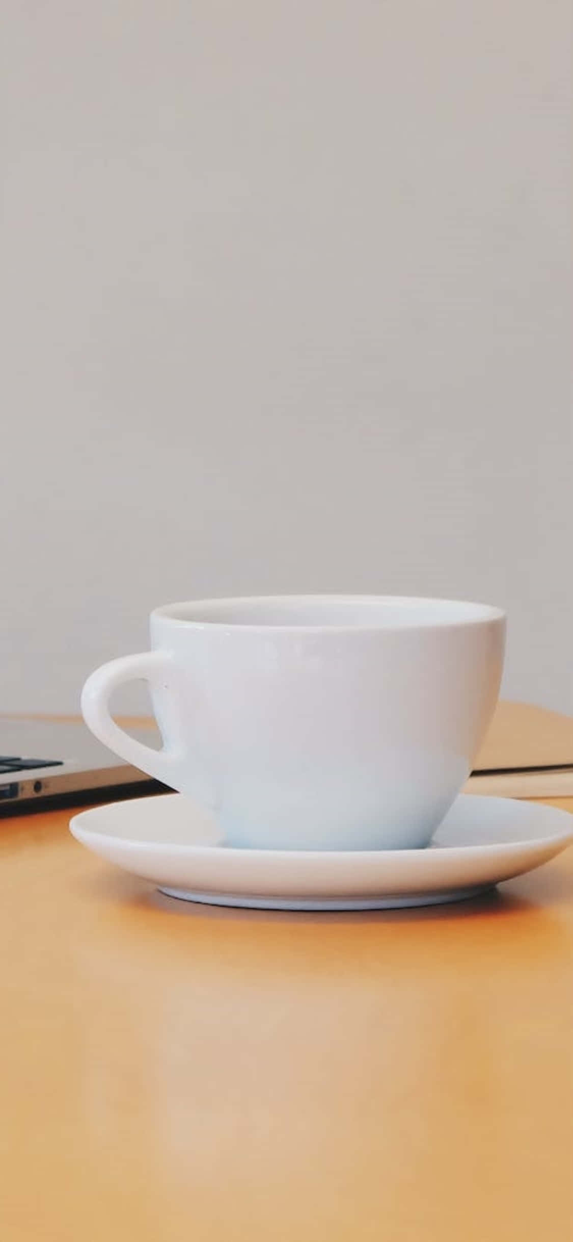 iPhone X 桌面背景：白色咖啡杯 壁紙 — 一個簡約的白色咖啡杯放在碟子上，旁邊有一部手提電腦在溫暖的木桌上。