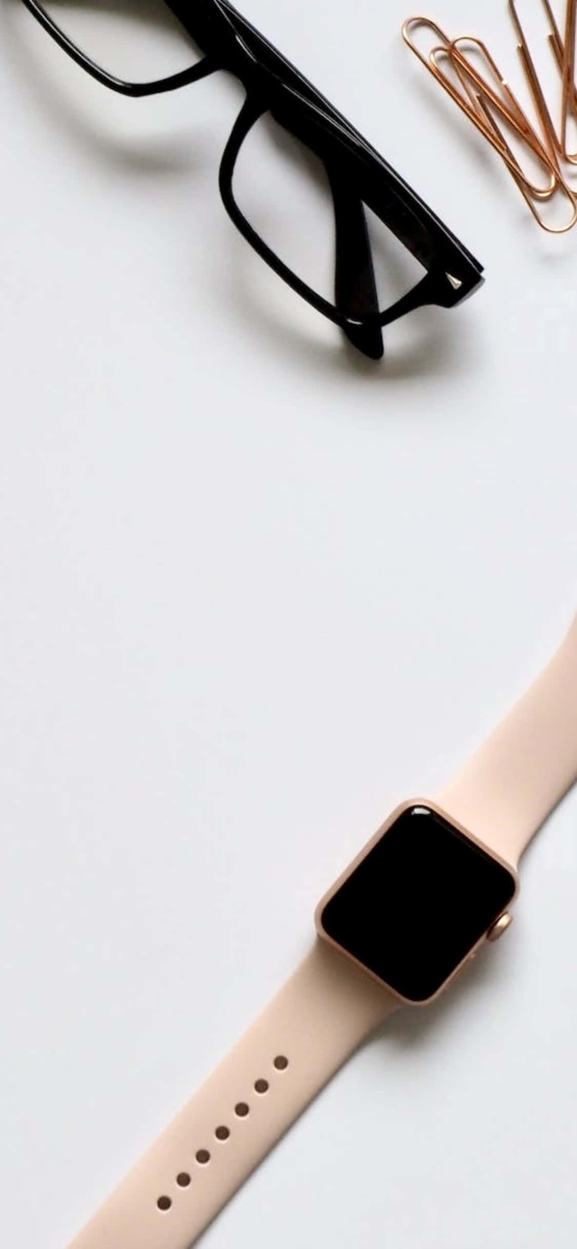 iPhone X 桌面背景：眼鏡與 Apple Watch 壁紙 — 極簡平鋪擺拍：黑色眼鏡、玫瑰金萬字夾和淡粉紅色智能手錶，置於純白表面上。