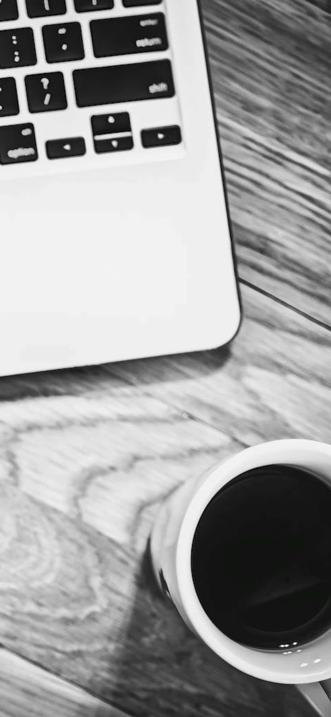 iPhone X 桌面背景：黑白照片（杯與筆記本電腦） 壁紙 — 一個單色特寫，顯示在簡約工作空間的木桌上有一部手提電腦和一杯咖啡。