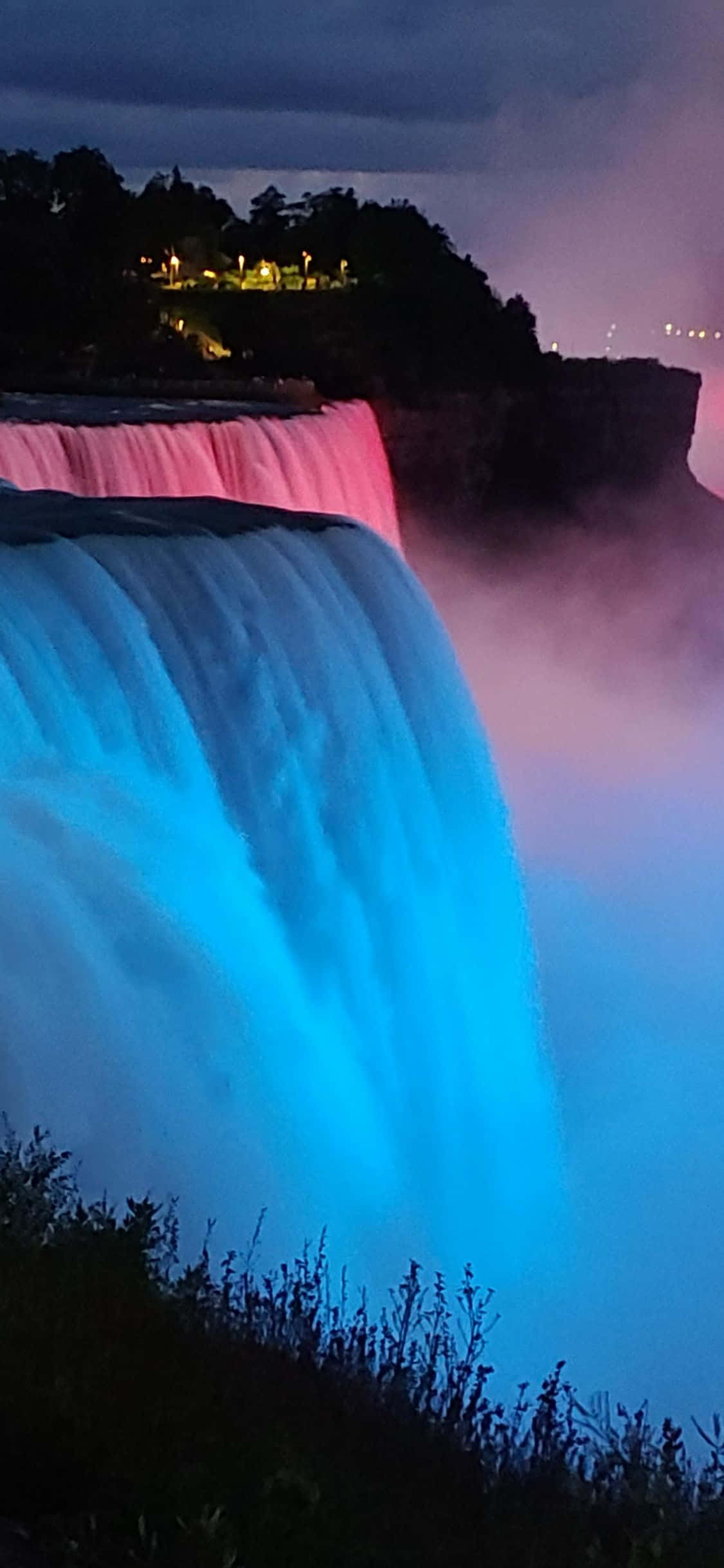 iPhone X 藍光照明尼亞加拉瀑布背景 壁紙 — 一座雄偉的瀑布在夜間被鮮豔的藍色和粉紅色燈光照亮，壯觀地傾瀉而下，薄霧在其底部旋轉。