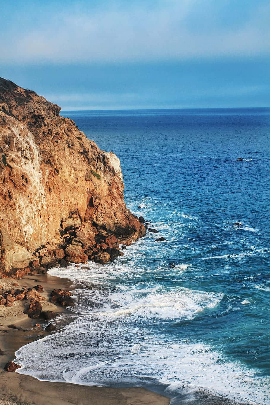 iPhone X 馬利布背景：寧靜海灘 壁紙 — 崎嶇的懸崖與在清澈藍天下拍打沙灘的碧綠海浪相遇。