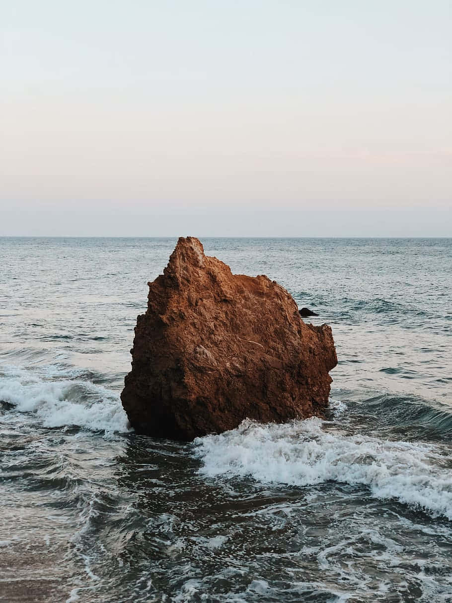 iPhone X 馬利布背景：巨石與海浪拍岸 壁紙 — 一座孤獨的紅褐色海蝕柱在暮色中傲然矗立於海洋中，溫柔的海浪拍打著它的底部。