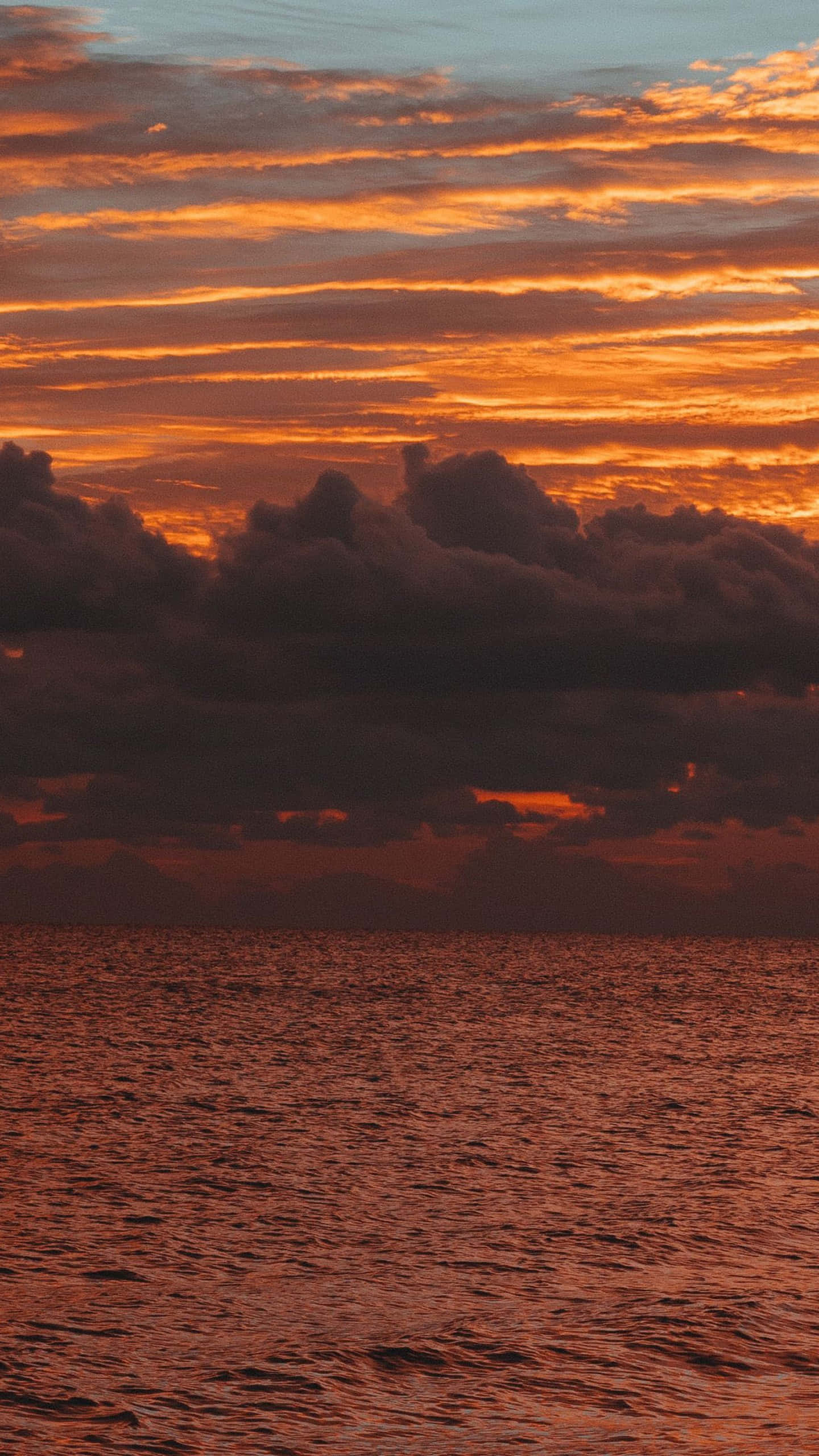 iPhone X 馬利布背景：暗雲日落 壁紙 — 熾熱的橙色夕陽用溫暖色調塗抹天空和海洋，下方烏雲密佈。