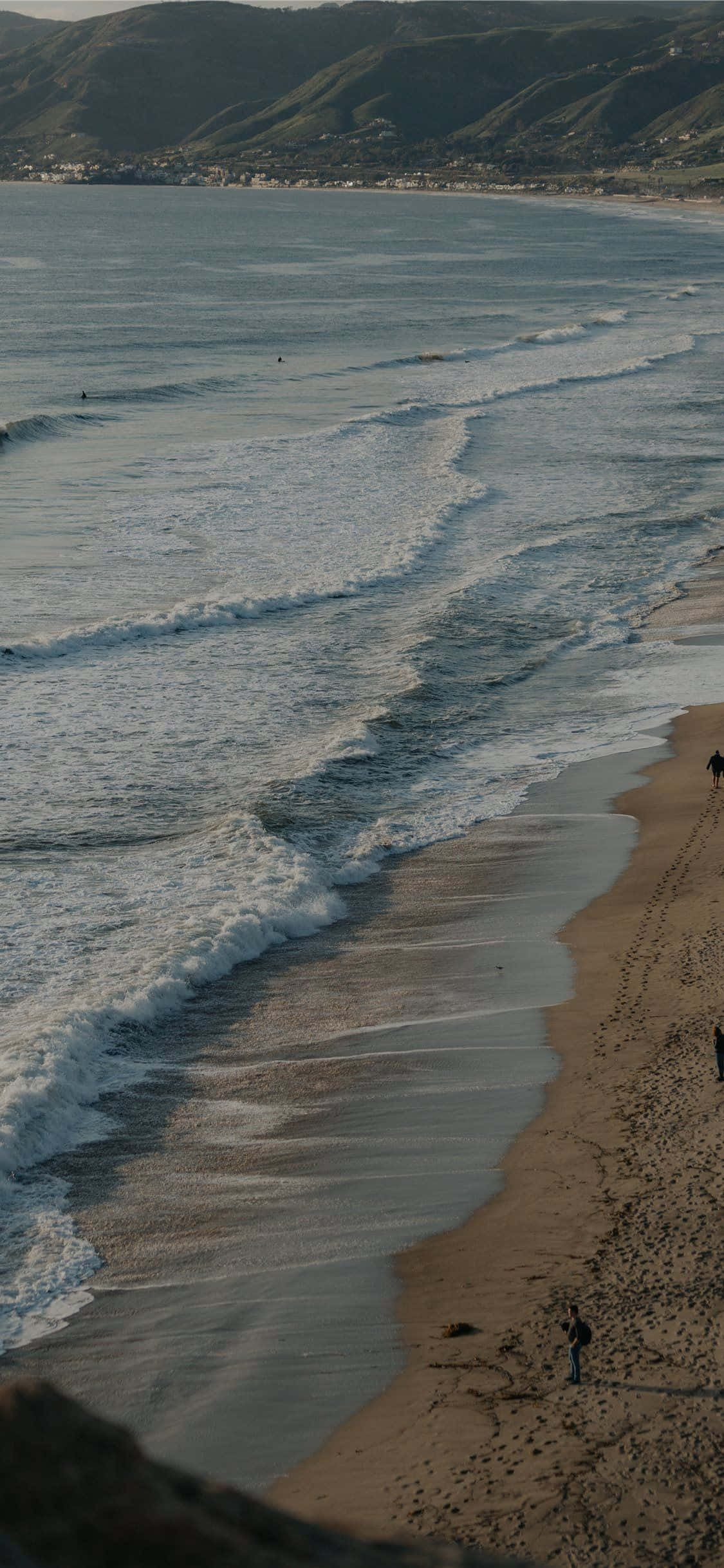 iPhone X 馬利布背景 泡沫海洋波浪 壁紙 — 海浪湧上沙灘，遠處山丘在柔和的金色光芒下。