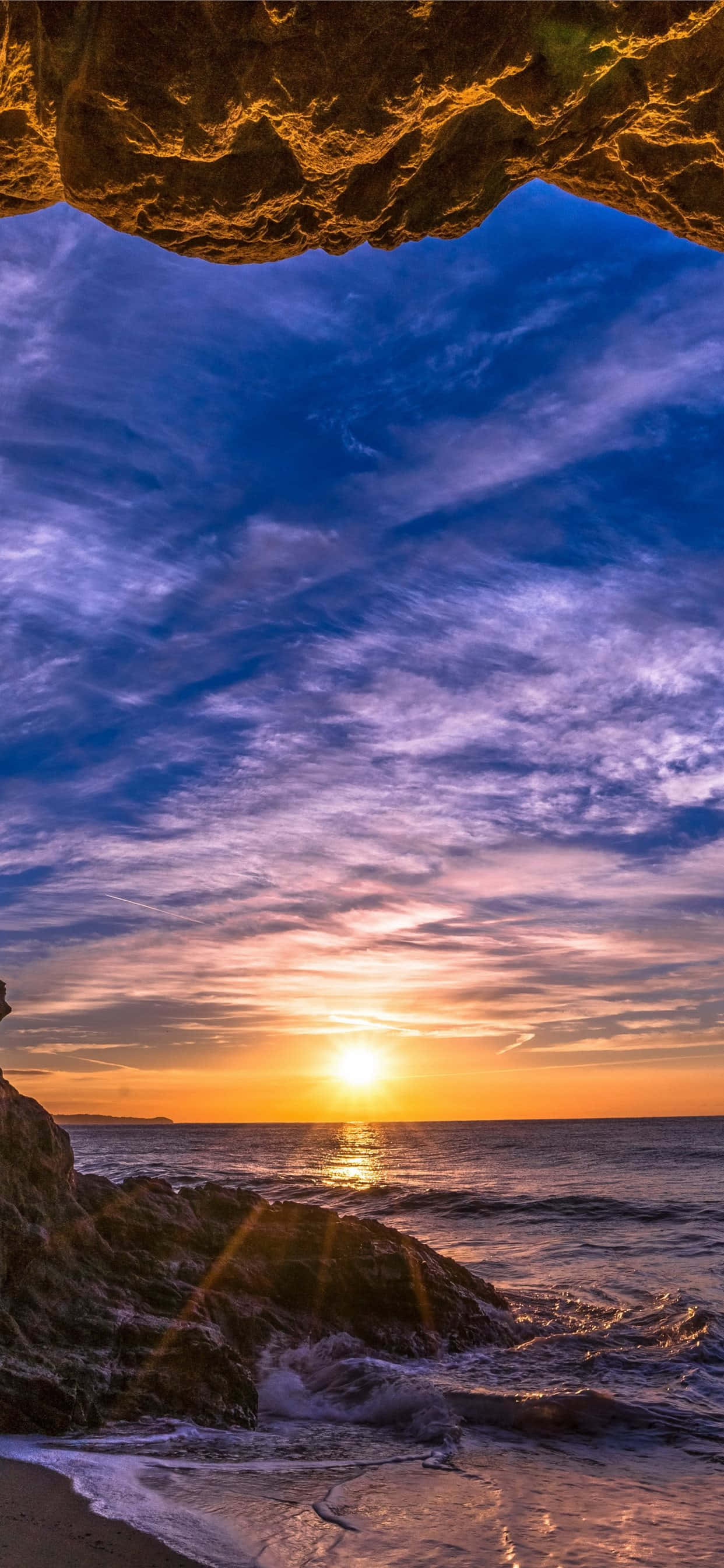 iPhone X 馬利布背景：洞穴觀海 壁紙 — 從岩石洞穴開口觀看的日落，金色光芒照亮雲層和下方拍打的波浪。