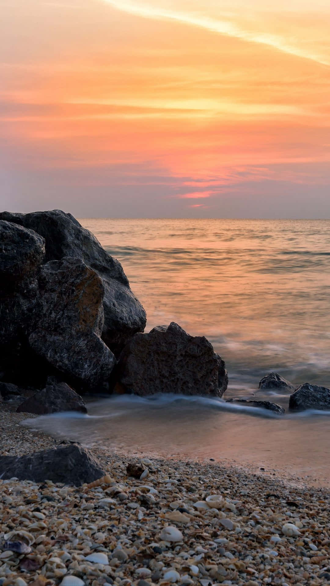 iPhone X 馬利布背景 海岸日落 壁紙 — 夕陽將天空染成火橙色，溫柔的海浪拍打著卵石海灘上的暗色岩石。
