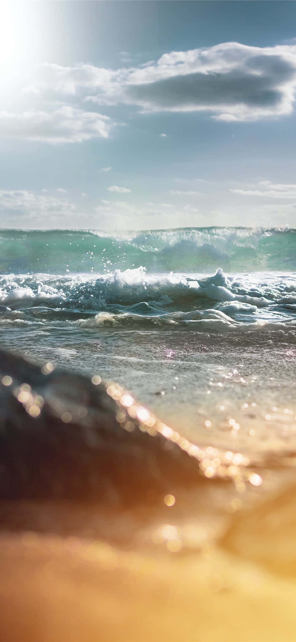 iPhone X 馬利布背景 海浪低角度視角 壁紙 — 陽光照耀的海浪拍打沙灘，海水閃爍，前景有柔和的金色散景。