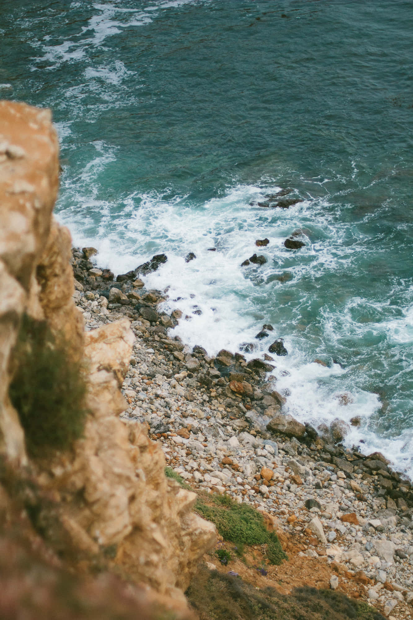 iPhone X 馬利布背景 海浪拍擊懸崖 壁紙 — 從高處俯瞰，碧綠色的波浪拍打著崎嶇的懸崖和卵石海灘。