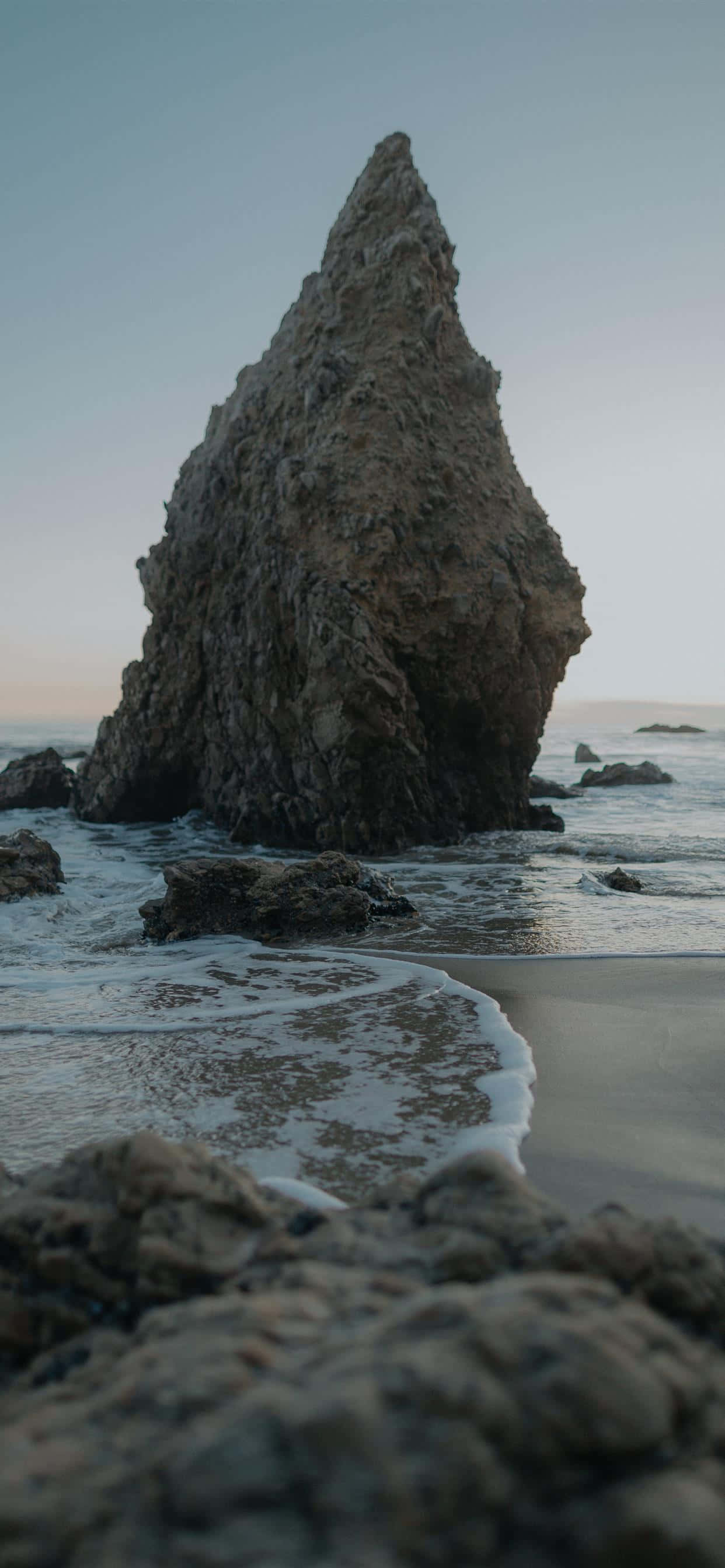 iPhone X 馬利布背景：艾爾馬塔多州立海灘 壁紙 — 一座高聳崎嶇的岩石形成孤獨地矗立在霧氣瀰漫的海灘上，溫柔的海浪在黃昏時分拍打著它的底部。
