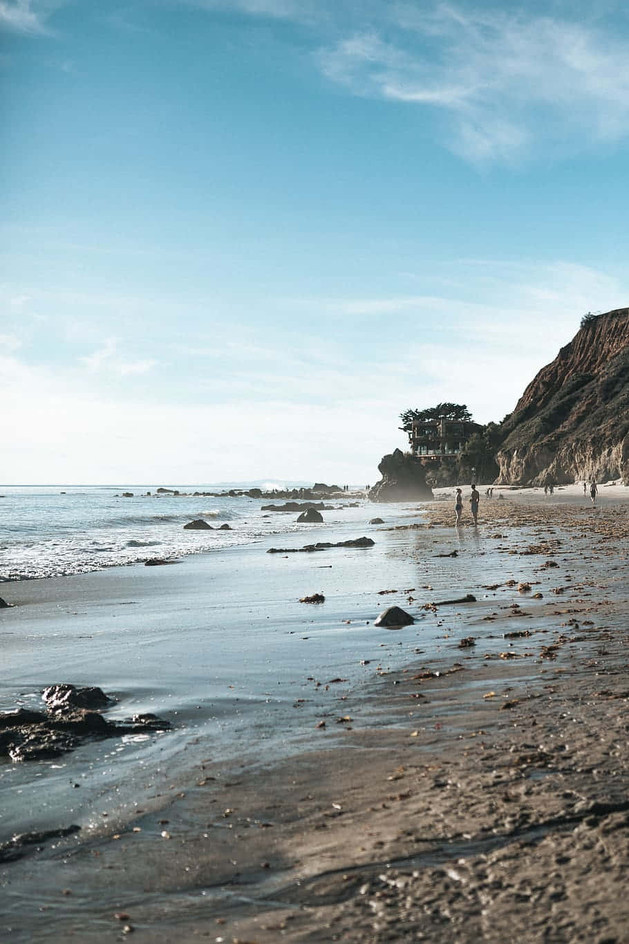 iPhone X 馬里布背景：岩石海灘岸邊 壁紙 — 寧靜的海灘場景，溫柔的海浪拍打濕沙，藍天清澈，遠處有懸崖，遊客稀疏。