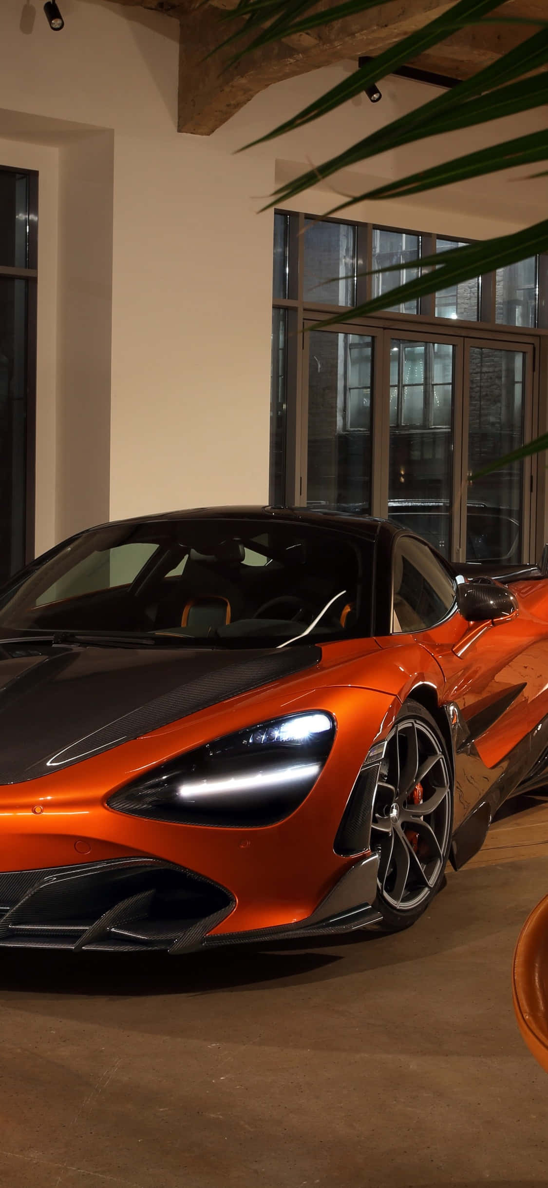 iPhone X 麥拉倫 720S 橙色背景 壁紙 — 一輛時尚的橙色和黑色McLaren跑車在溫暖的室內燈光下，旁邊有大窗戶和綠色棕櫚葉，閃閃發光。
