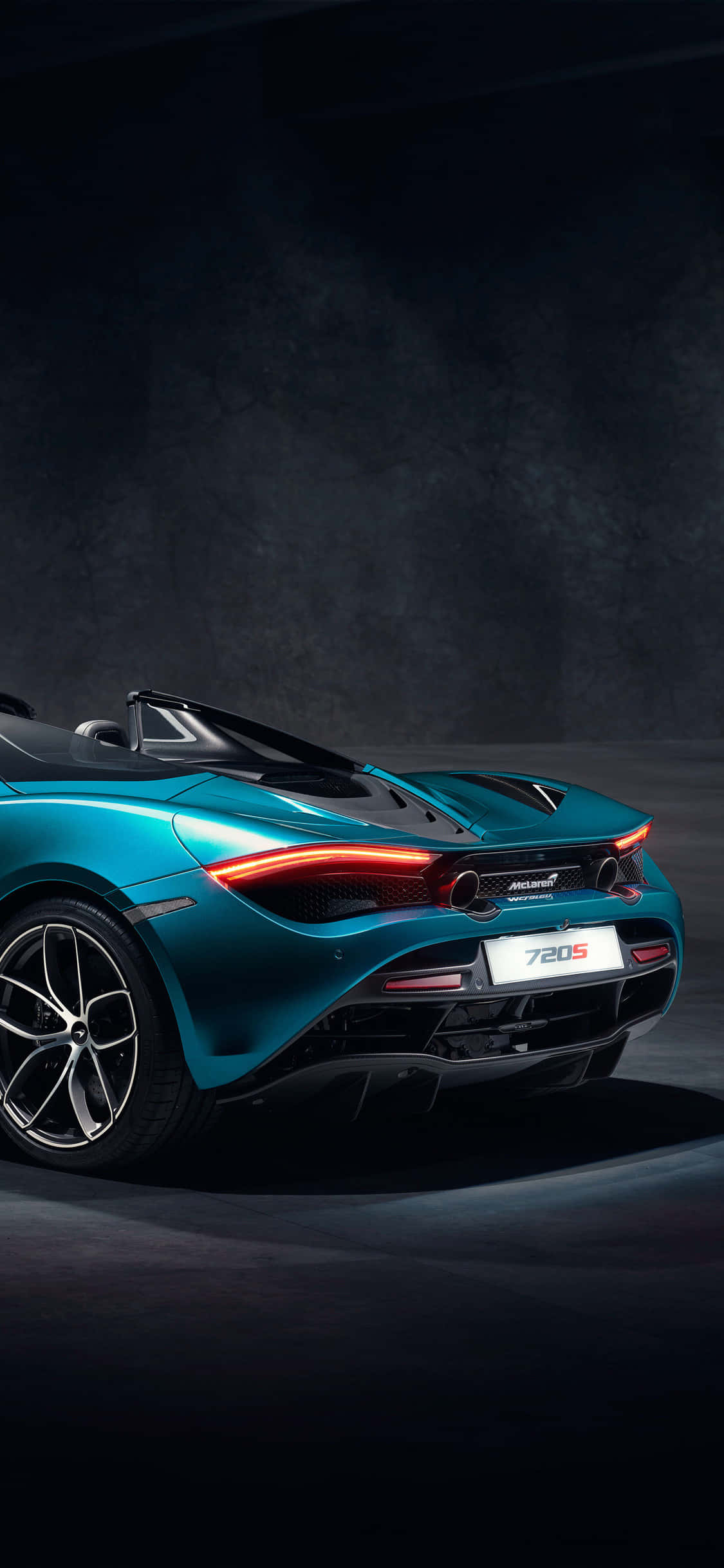 iPhone X 麥拉倫 720S Spider Pista 背景 壁紙 — 一架時尚的青色McLaren 720S Spider開篷車在戲劇性的影樓燈光下，在深色紋理牆壁前閃閃發光。