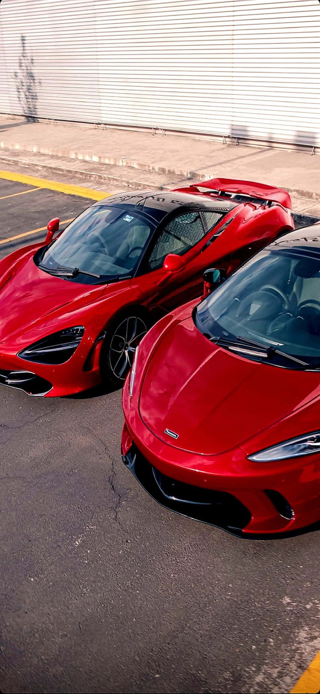 iPhone X 麥拉倫 720S 紅色車款背景 壁紙 — 兩輛亮面紅色McLaren超級跑車並排停放在陽光明媚的城市空地，背景是波紋金屬牆。