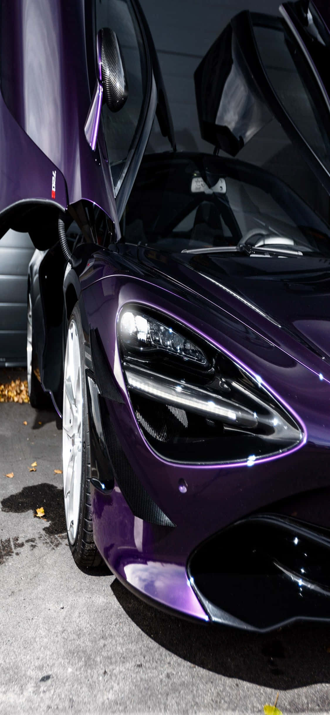 iPhone X 麥拉倫 720S 紫色車門開啟背景 壁紙 — 一架亮紫色McLaren跑車，剪刀門打開，停在濕漉漉的路面上，周圍散落著秋天的落葉。