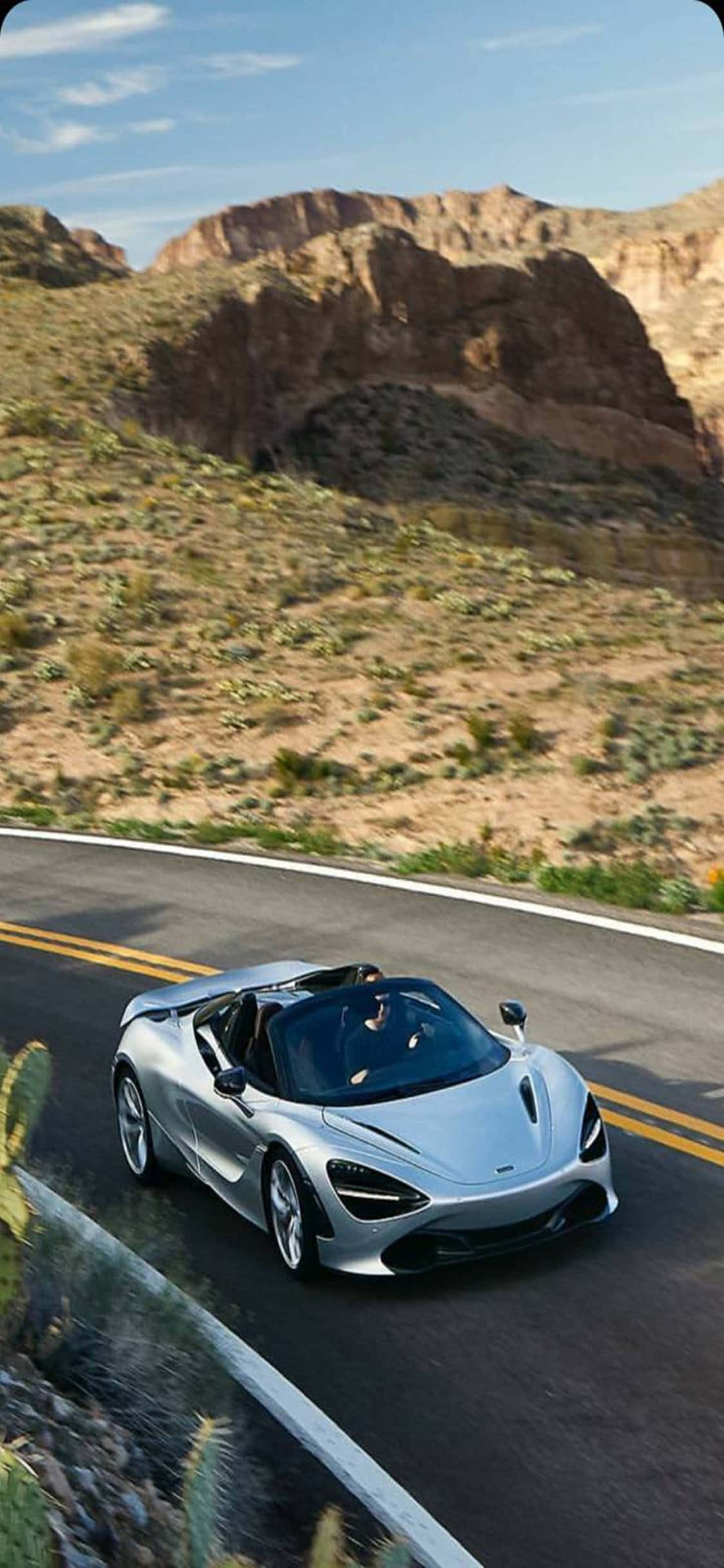 iPhone X 麥拉倫 720S Spider 荒漠駕駛背景 壁紙 — 一輛銀色McLaren 720S Spider在晴朗藍天下，沿著兩旁有岩石峭壁和仙人掌的沙漠公路巡航。