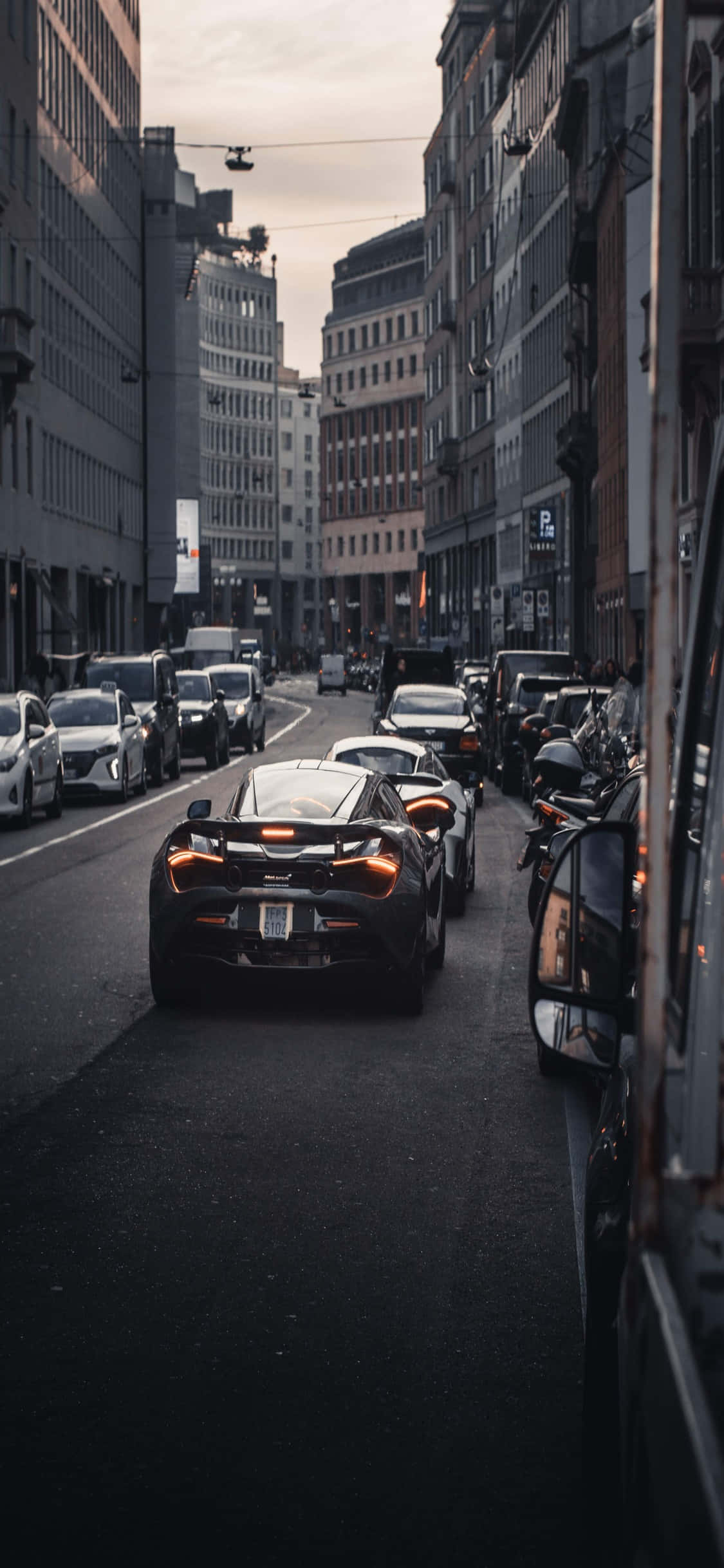 iPhone X 麥拉倫 720S 城市景觀背景 壁紙 — 一輛時尚黑色跑車在暮色蒼茫的天空下，行駛在兩旁高樓林立的狹窄城市街道上。