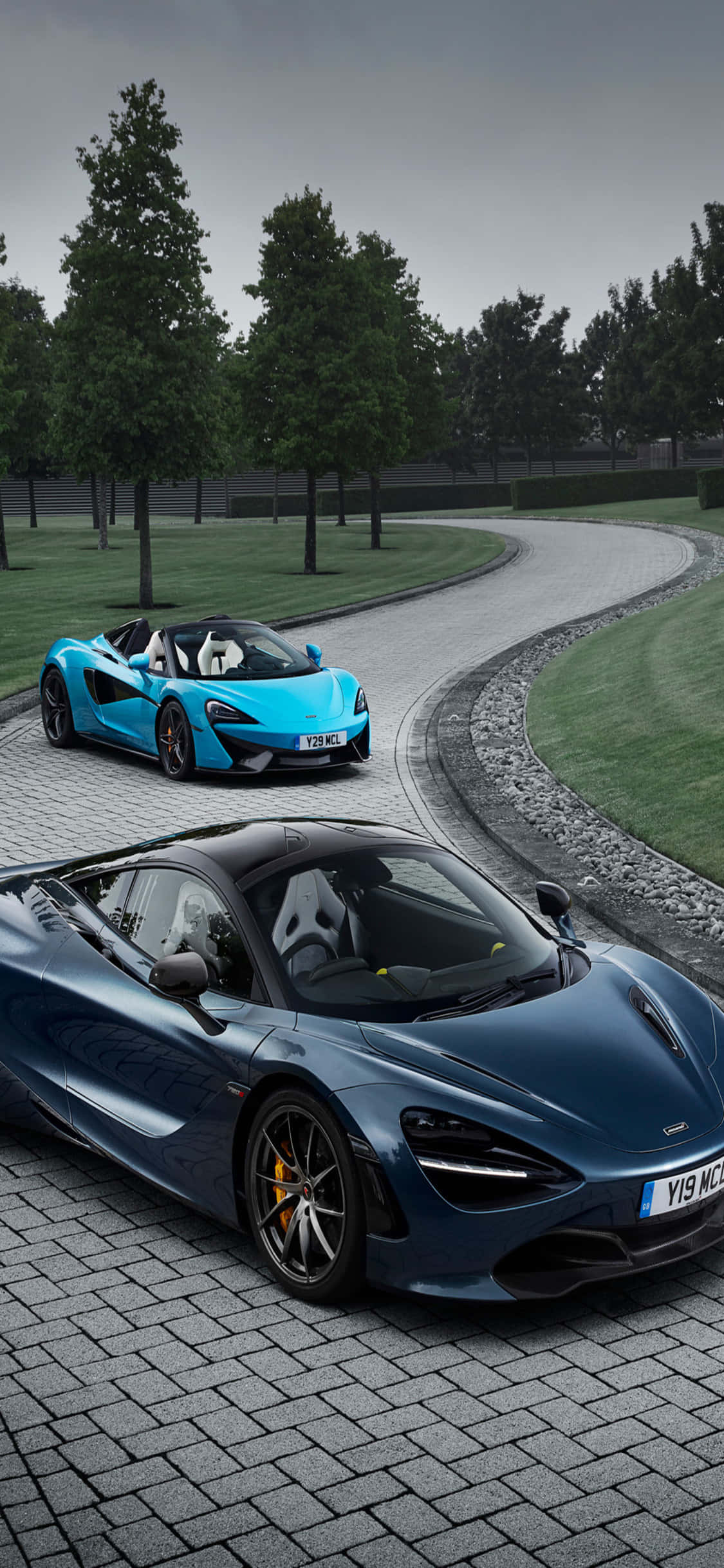 iPhone X 麥拉倫 720S 背景 壁紙 — 兩輛時尚的McLaren超級跑車，一輛藍色一輛深藍色，停泊在彎曲的石頭車道上，周圍綠樹成蔭，天空多雲。